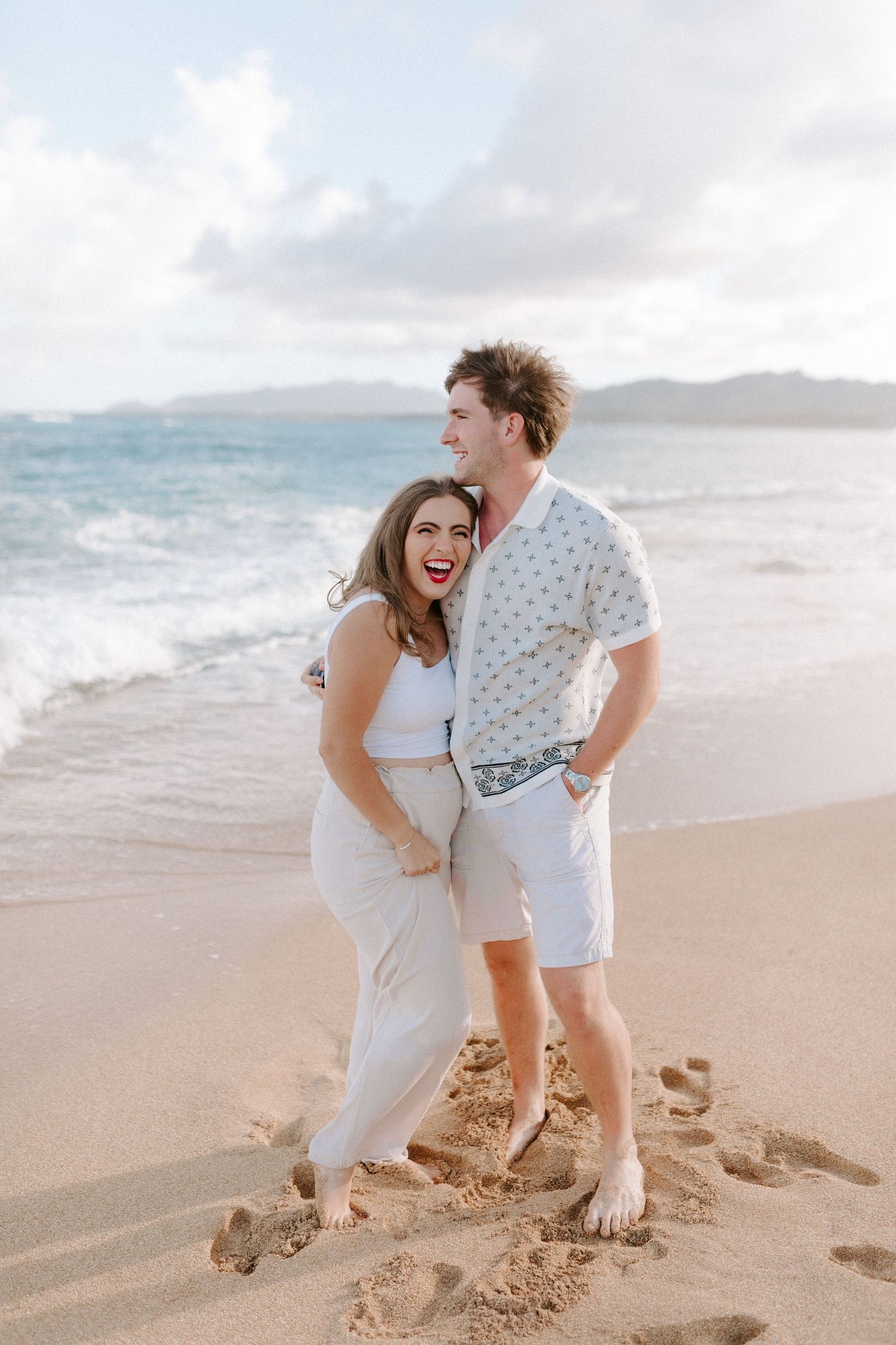 A couple shares a joyful moment together on a sandy beach with mountains and ocean waves in the background.