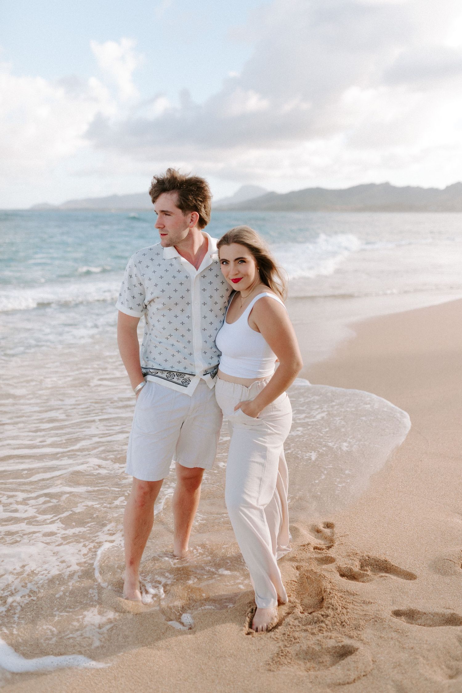 Two people pose together on a tropical beach wearing white and light colored summer clothing.