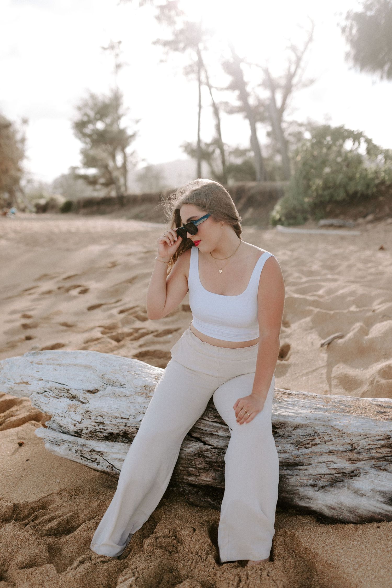 A person in all white clothing sits on driftwood at the beach taking photos.