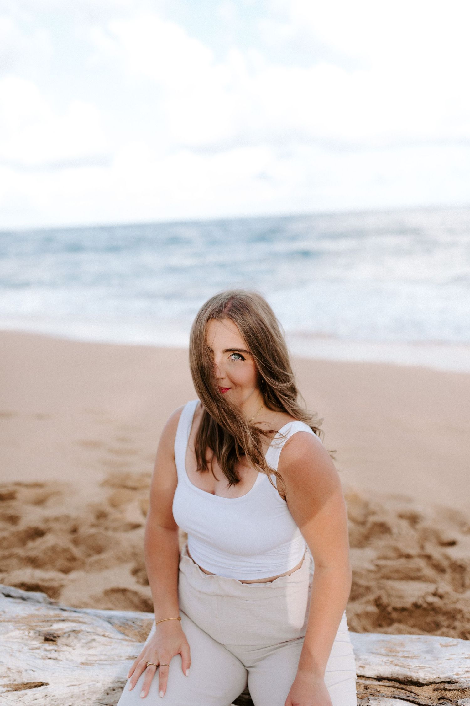 A casual beach portrait of someone sitting on driftwood in white summer attire.