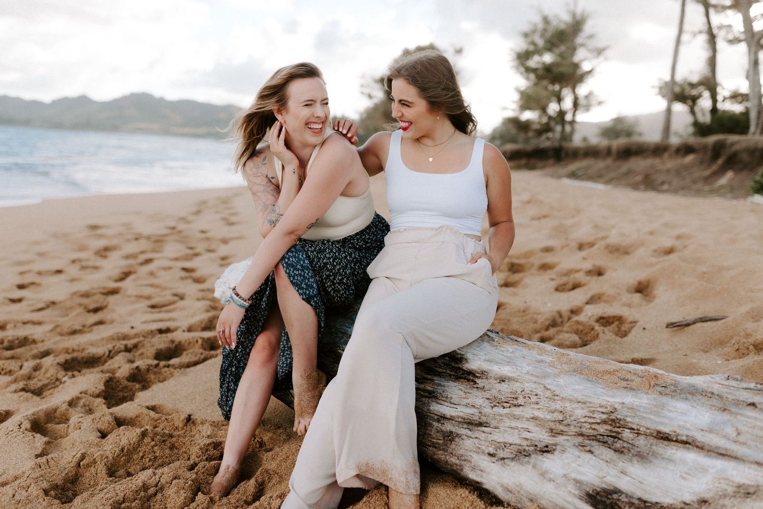 Two friends share a laugh while sitting together on driftwood at the beach.