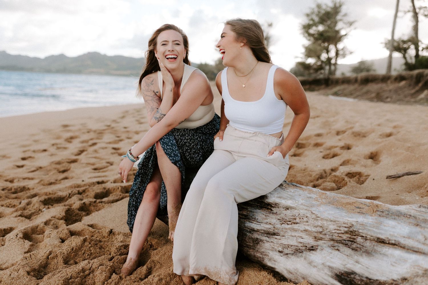 Two friends share genuine laughter while sitting on driftwood at a scenic beach.