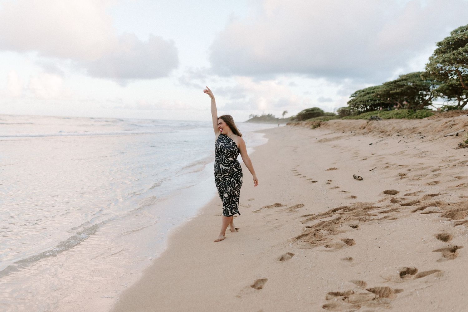 A person in a patterned dress dances joyfully along the shoreline at sunset.