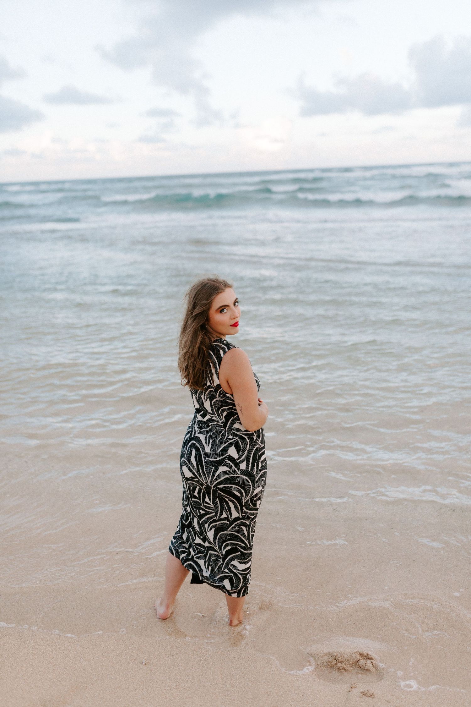Woman in a patterned dress walks barefoot along a serene beach at sunset.