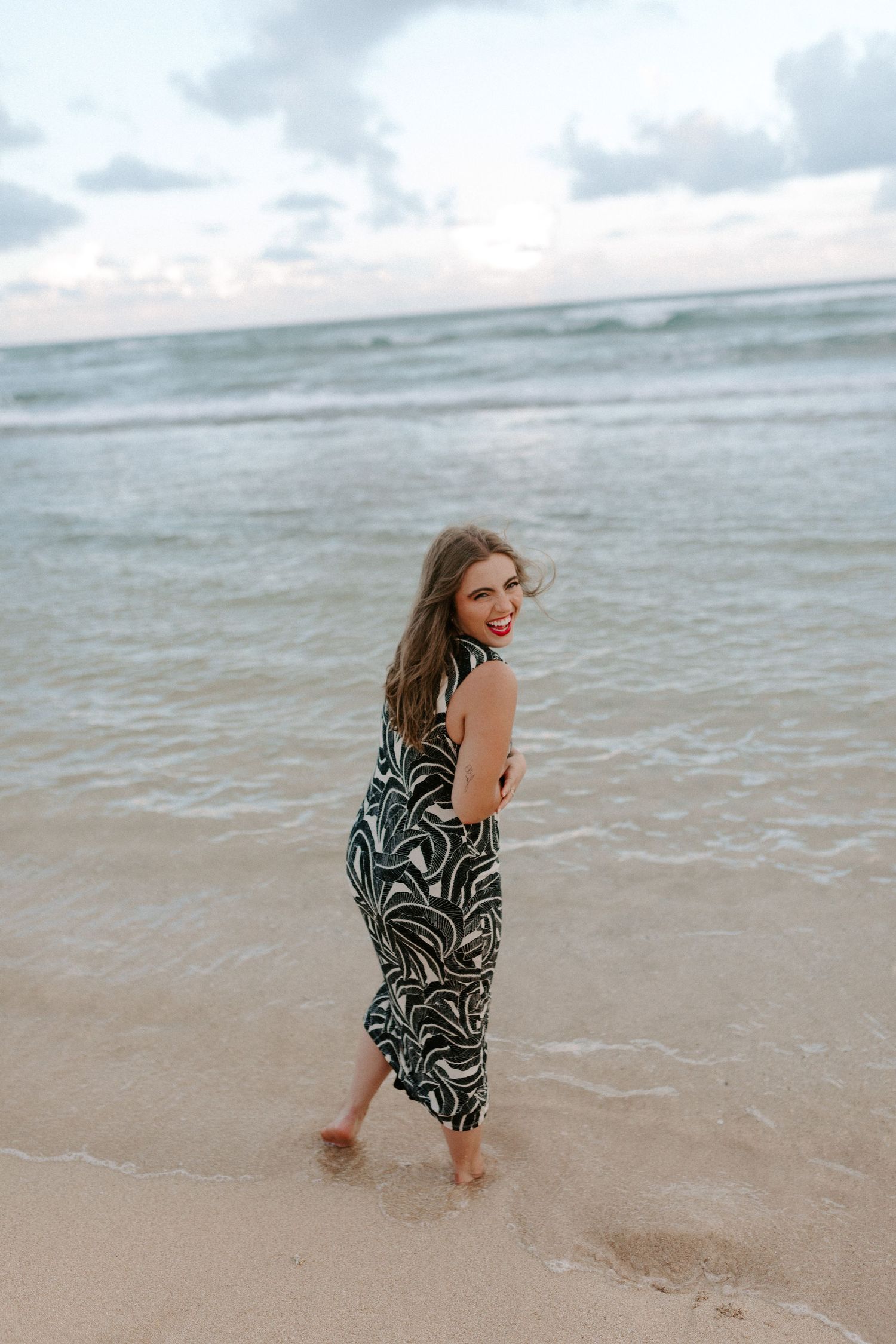 Figure wearing a black and white tropical print dress poses on a sandy shoreline.