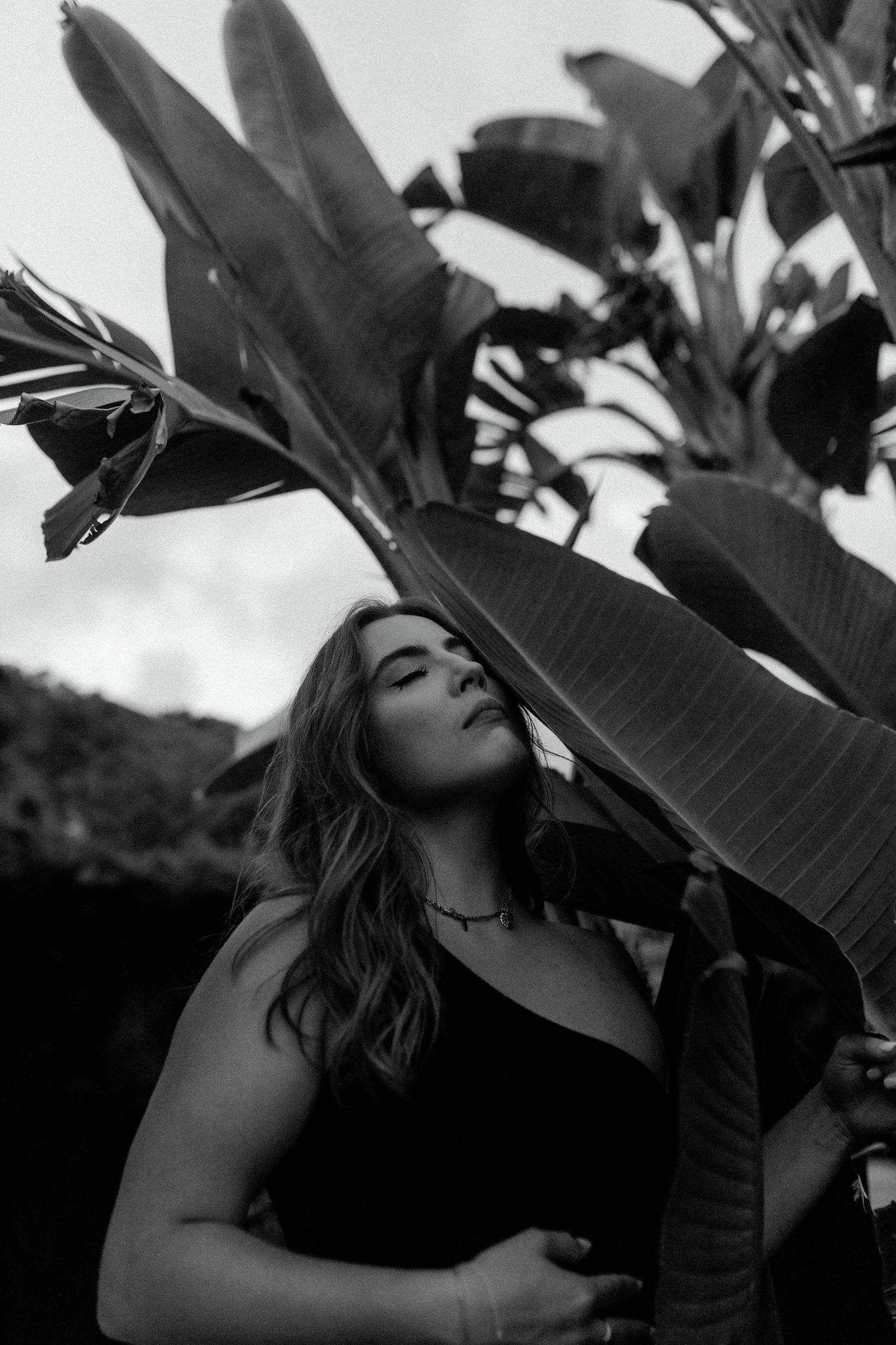 Artistic black and white portrait with large tropical leaves in the foreground.