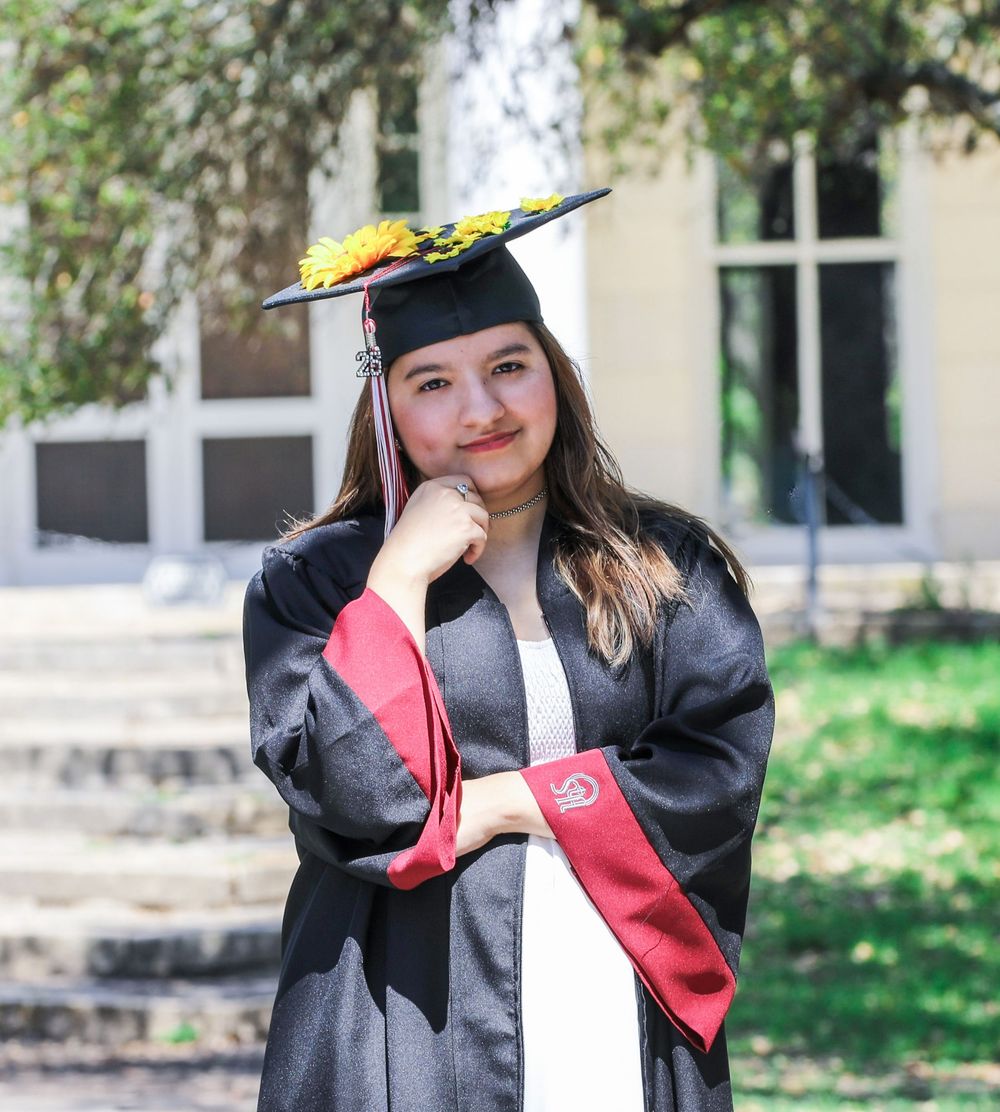 Graduate in traditional black cap and gown with yellow decoration poses outdoors on campus steps.
