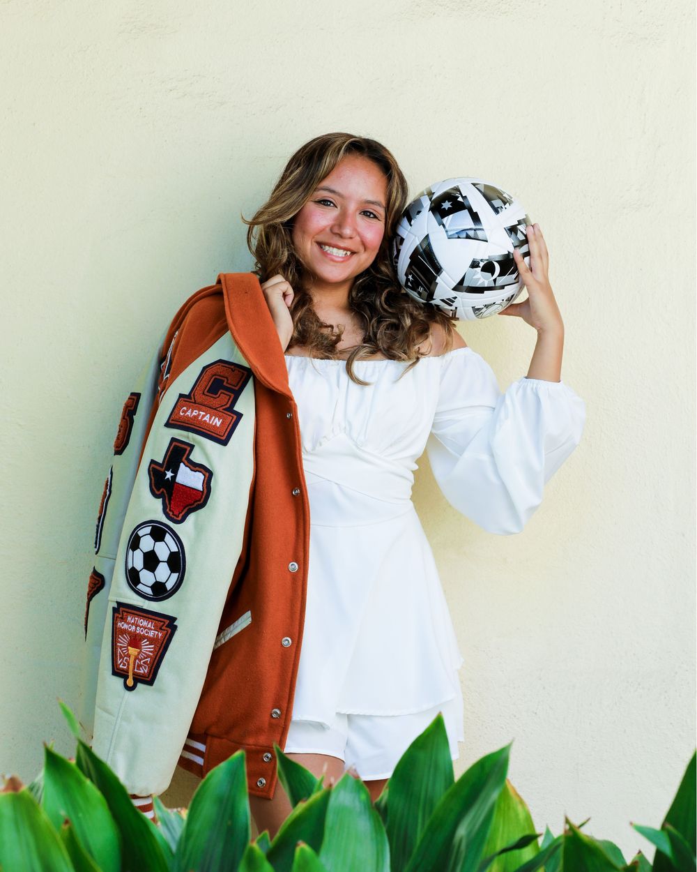 Person in white sundress poses with a soccer ball and athletic varsity jacket decorated with sports patches against beige wall.