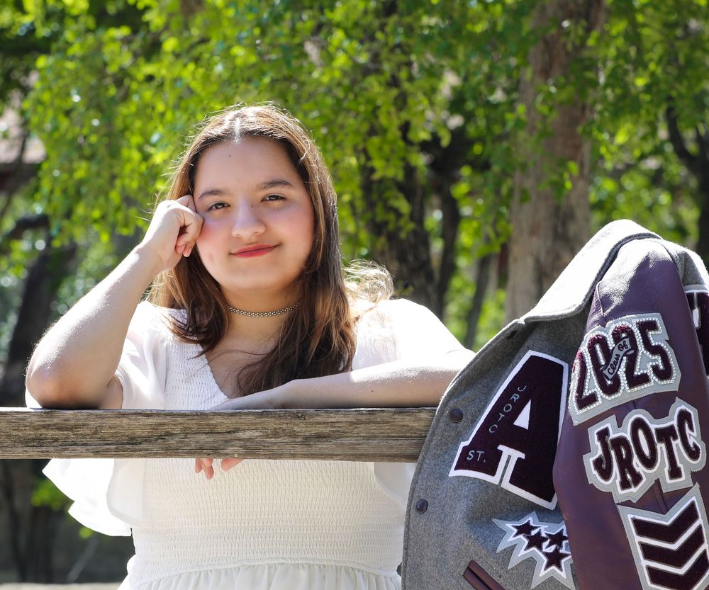 Student in white t-shirt poses with varsity letterman jacket in outdoor park setting during sunny day.