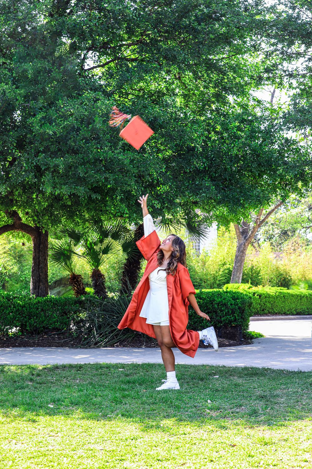 Graduate in red cap and gown throws graduation cap into air on sunny day in grassy park setting.