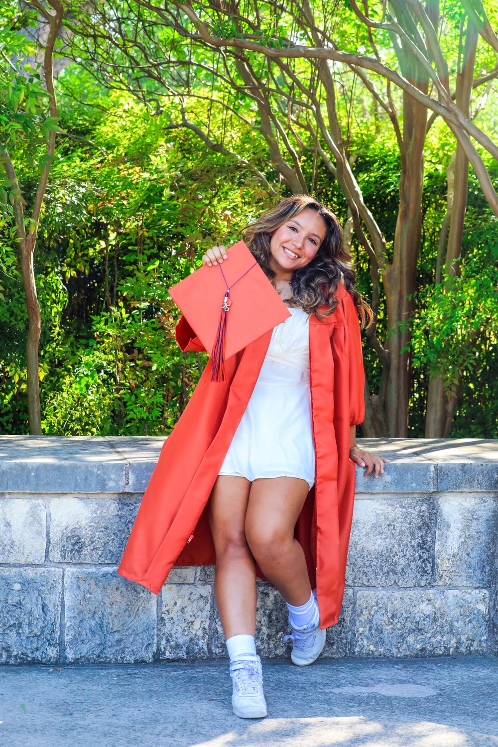 Graduate in bright red commencement gown poses cheerfully on stone steps surrounded by lush green foliage.
