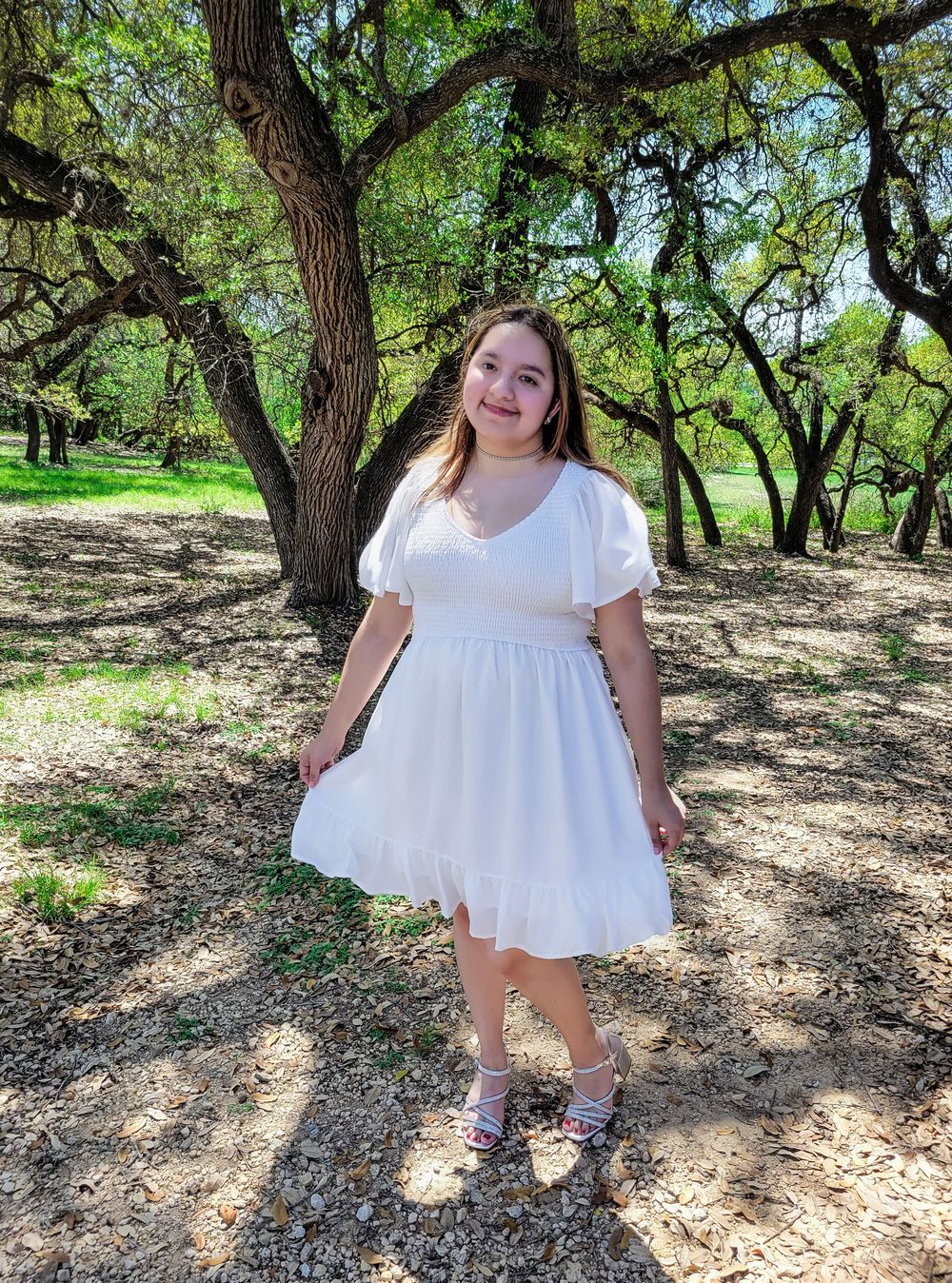 A person in a white summer dress poses outdoors in a park setting with oak trees and dappled sunlight in the background.