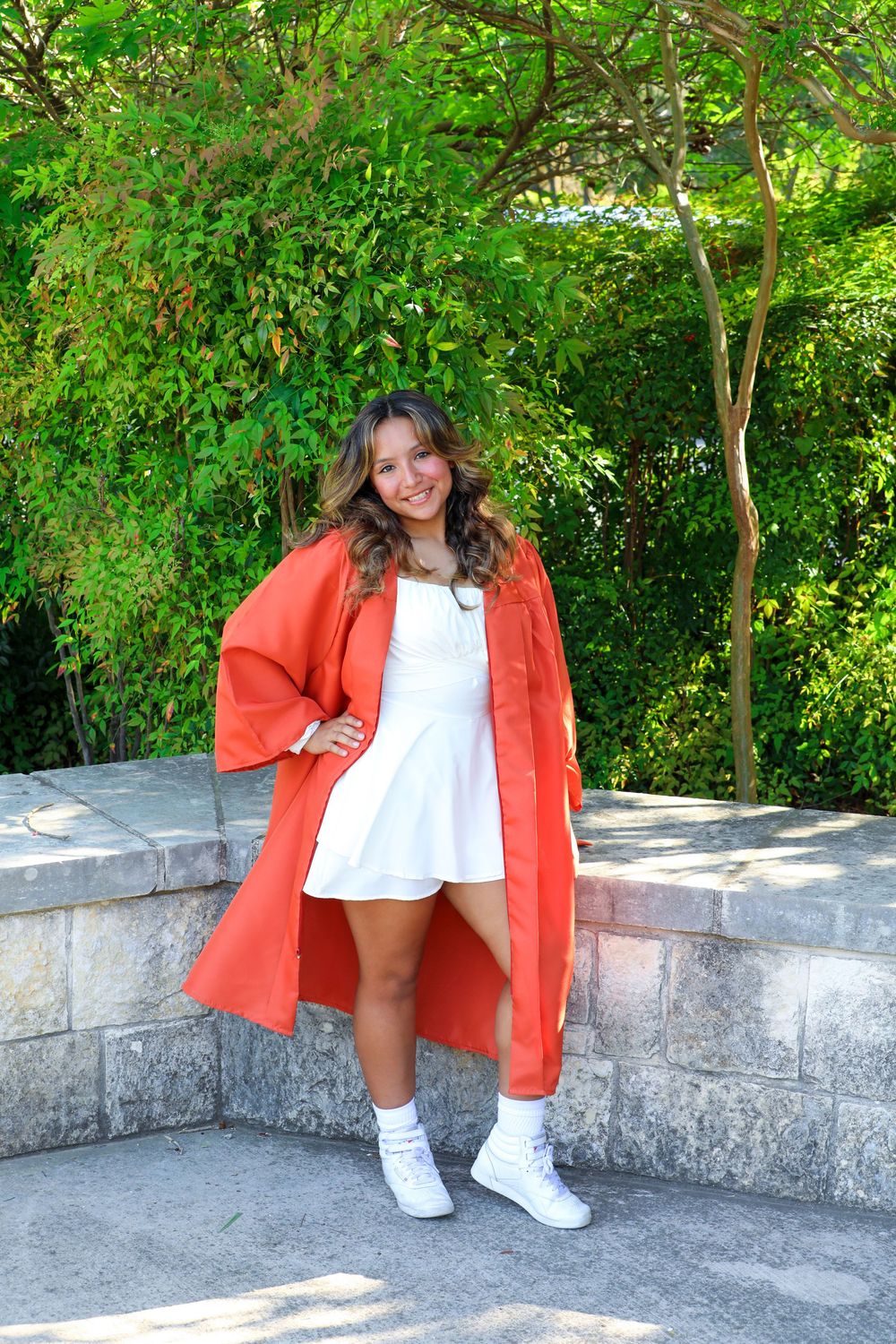 A person in a bright coral coat and white dress poses on stone steps surrounded by lush green foliage.