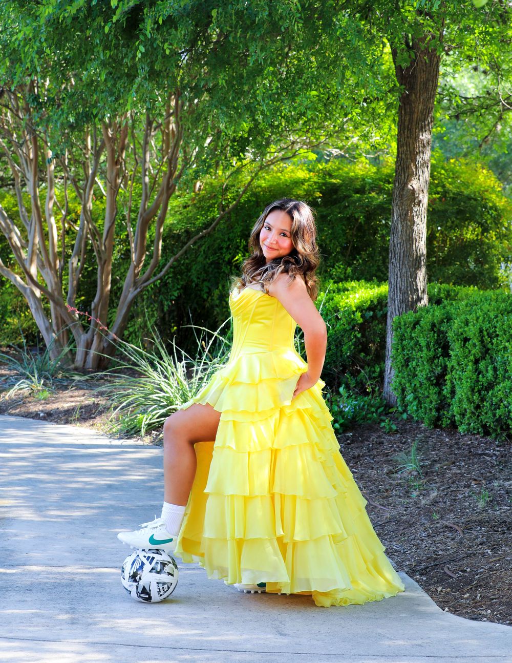 A bright yellow tiered formal gown is showcased outdoors next to a soccer ball on a garden pathway.