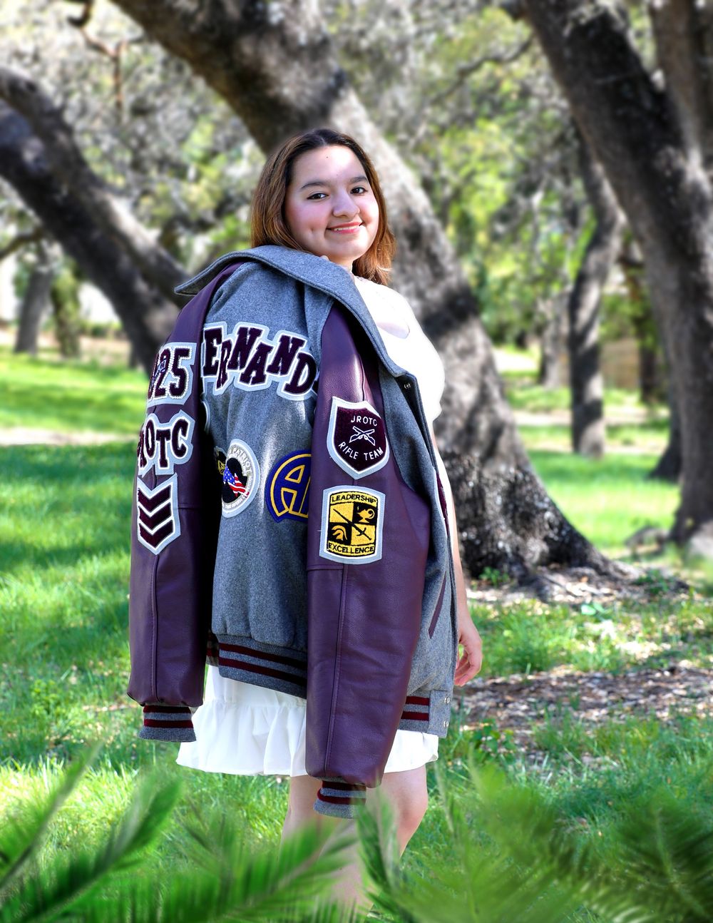 A student in a varsity letterman jacket decorated with patches poses outdoors in a grassy area with trees in the background.
