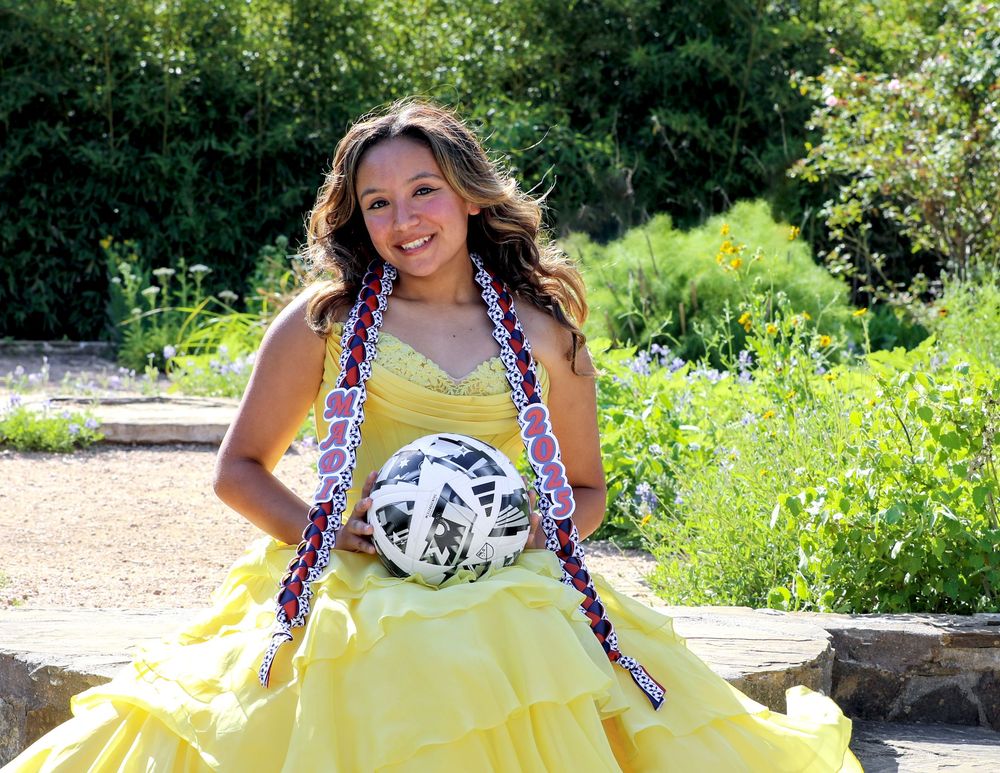 Someone wearing a bright yellow dress poses outdoors on a sunny day while holding a soccer ball.