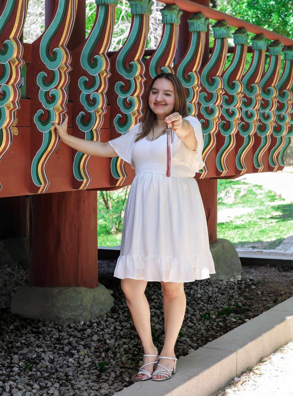 A person in a white summer dress poses next to ornate teal and brown architectural details in an outdoor setting.