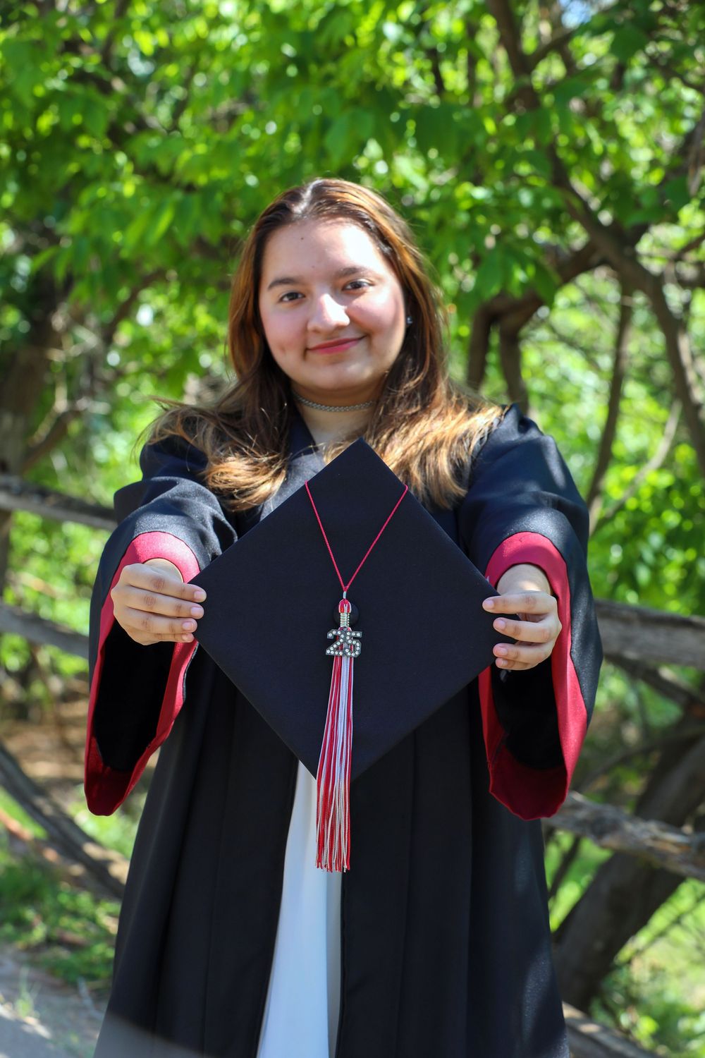Graduate in black cap and gown with red tassel holds up their graduation cap in front of green foliage outdoors.