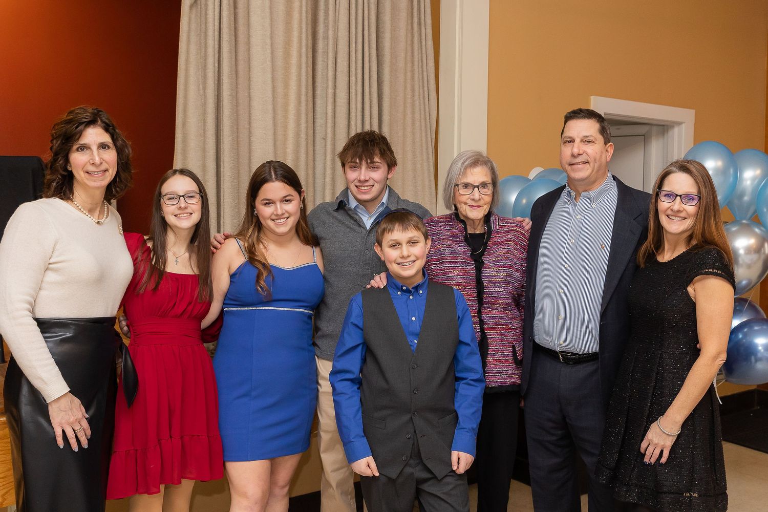 A group of people dressed up for a formal occasion pose together with blue balloons decorating the venue.