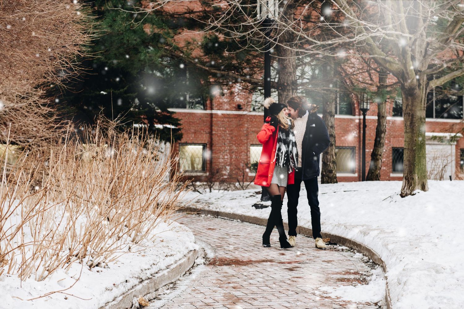 A couple strolling along a snow-covered brick pathway near ornate campus buildings. Massachusetts Engagement Photographer Serving New Englan