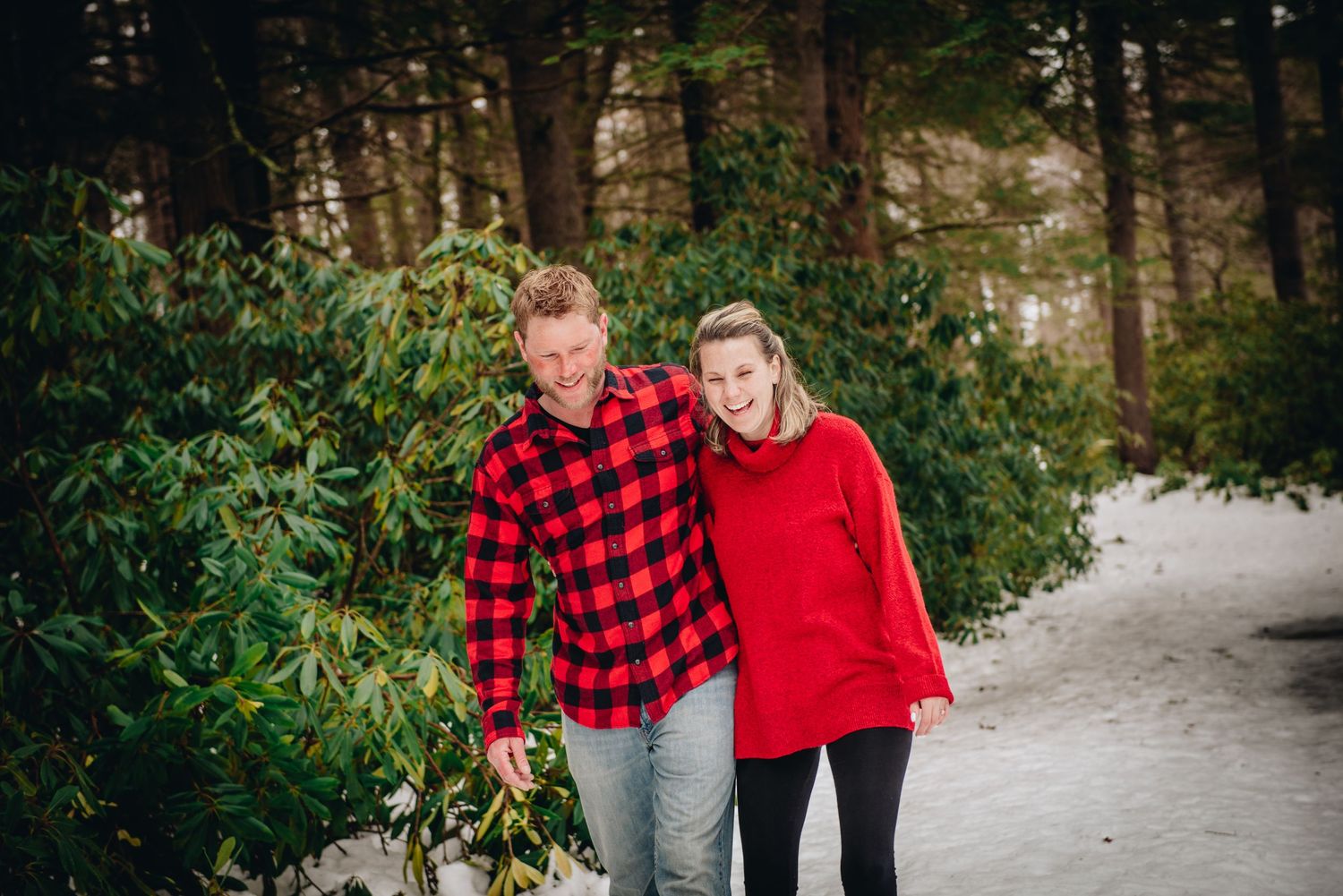A couple walking together on a snowy path in the woods wearing coordinating red outfits. Massachusetts Engagement Photographer- Affordable