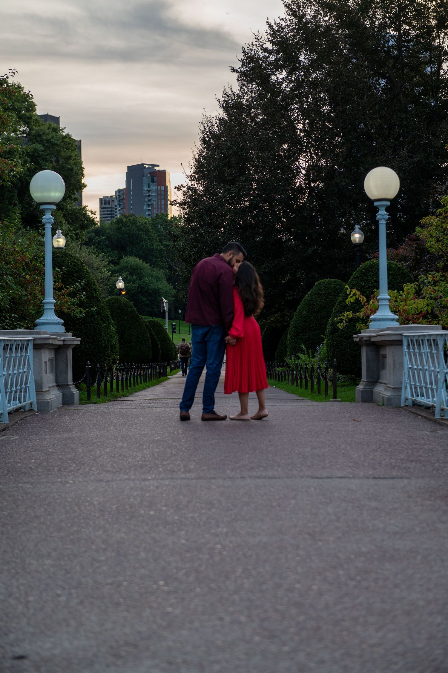 A couple shares an intimate moment on a bridge path at dusk with city lights in the background. Boston Engagement & Wedding Photography