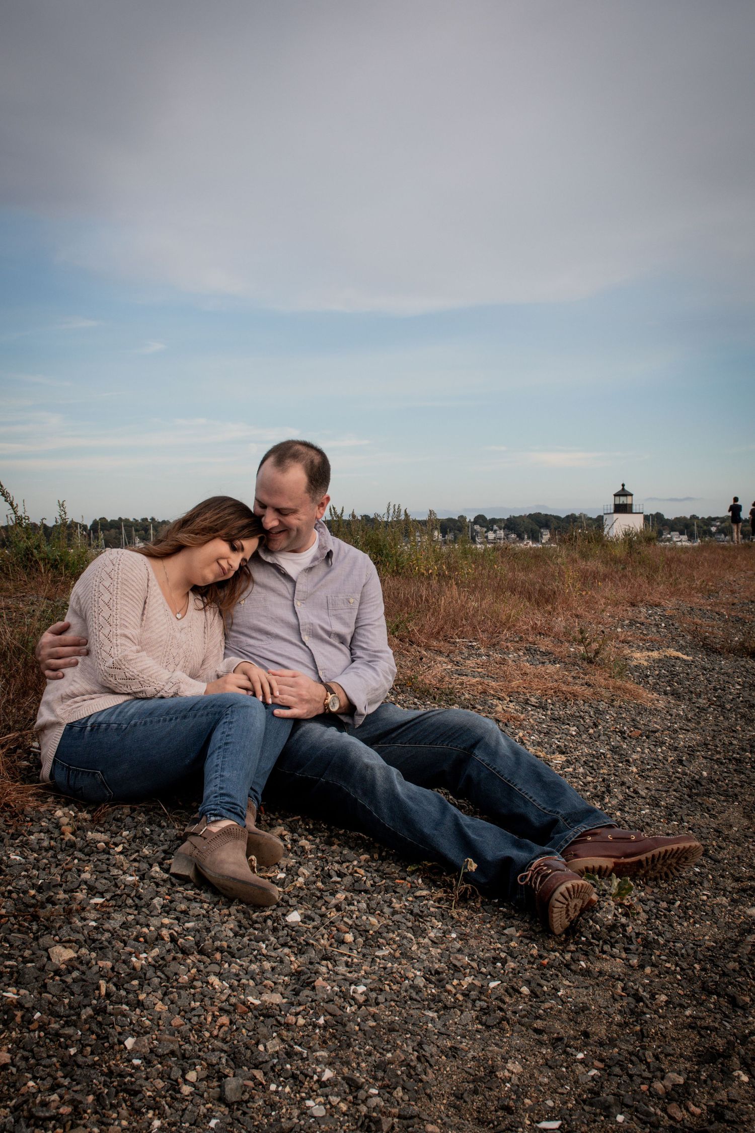 Intimate moment between couple resting on rocky shore with lighthouse in distance. Massachusetts Engagement Photographer Serving Salem, MA