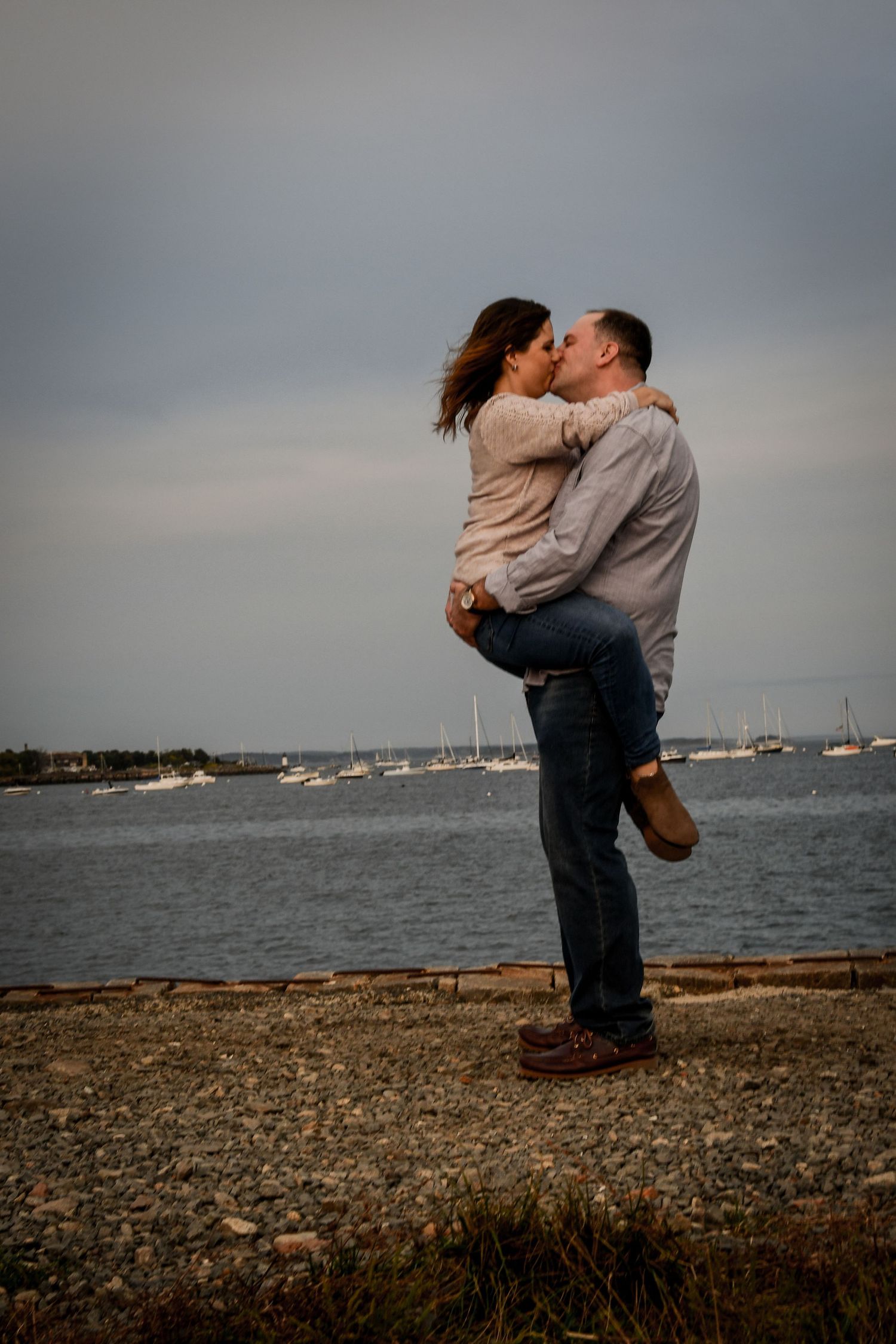 Romantic couple embracing on coastal path with boats and harbor in background. Massachusetts Engagement Photographer Serving New England