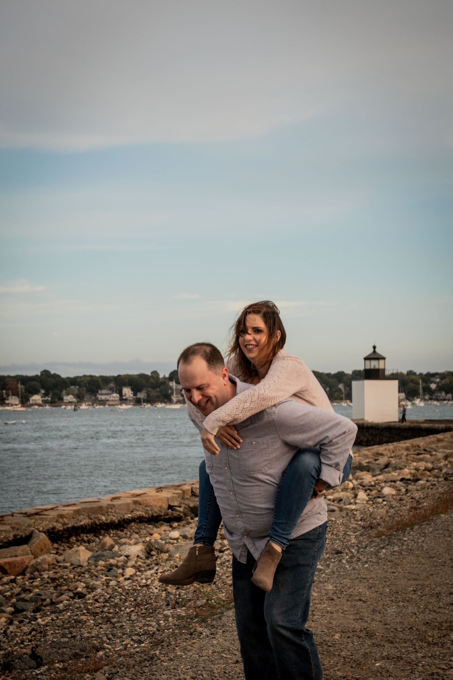 Playful piggyback ride near lighthouse with harbor view in background. Salem, Massachusetts Engagement Photographer Serving New England