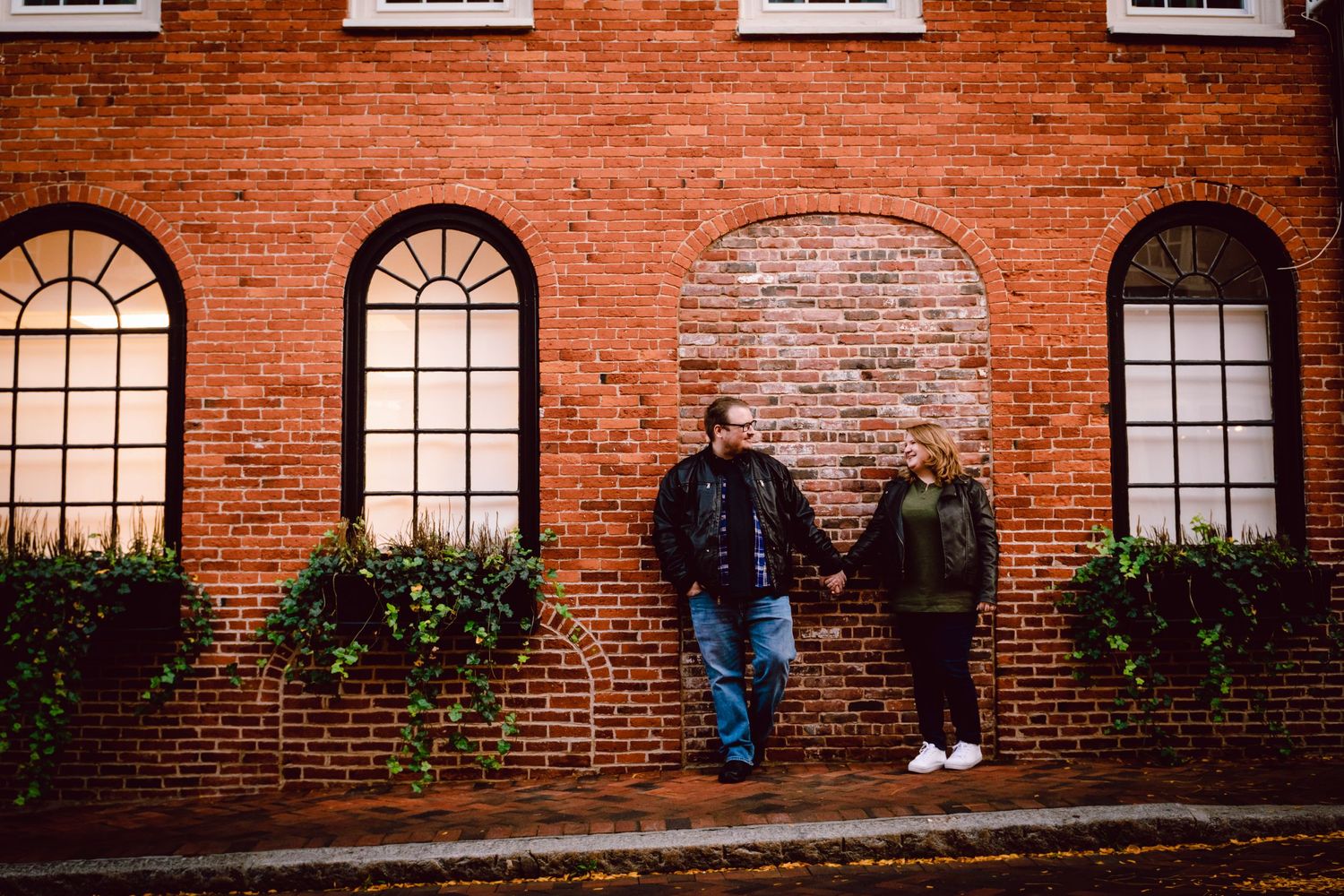 A couple stands against a historic brick building with arched windows. Massachusetts Engagement Photographer Serving Newburyport
