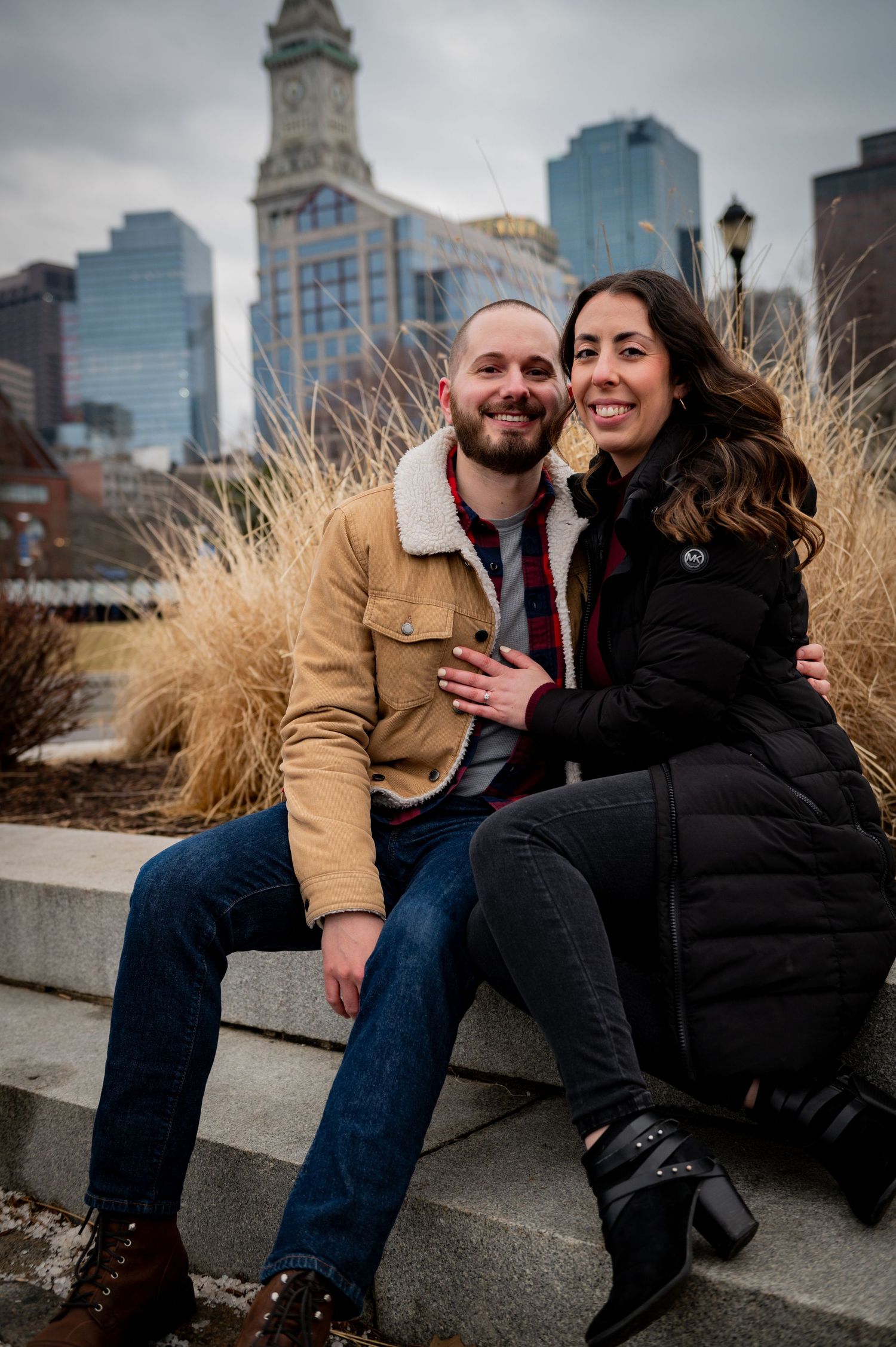 Couple sits together on concrete steps with Boston cityscape and Custom House Tower in background. Boston Engagement Photos- Affordable