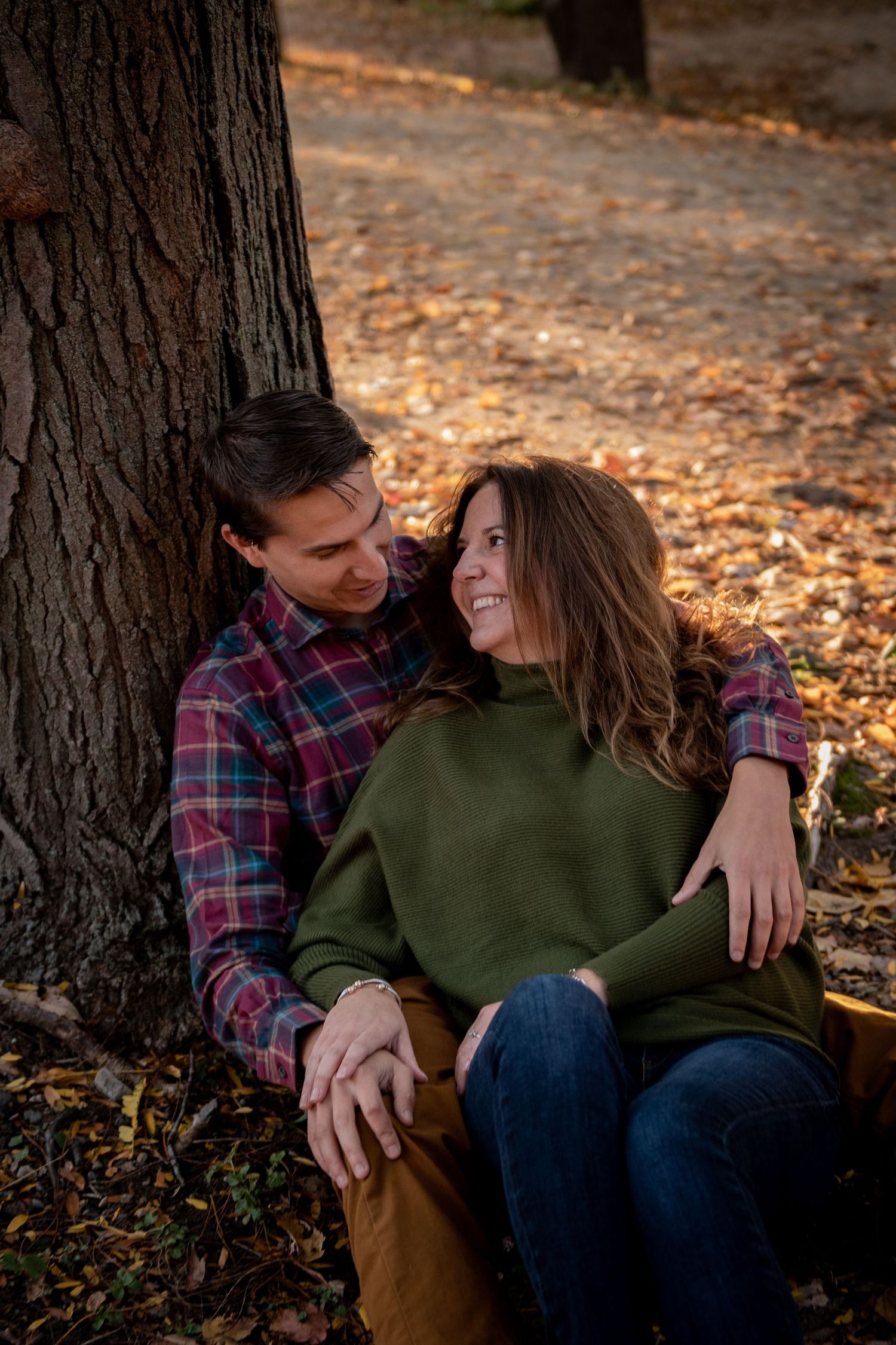 Romantic autumn engagement photo of couple cuddling against tree trunk surrounded by fallen leaves. Massachusetts Engagement Photographer Se