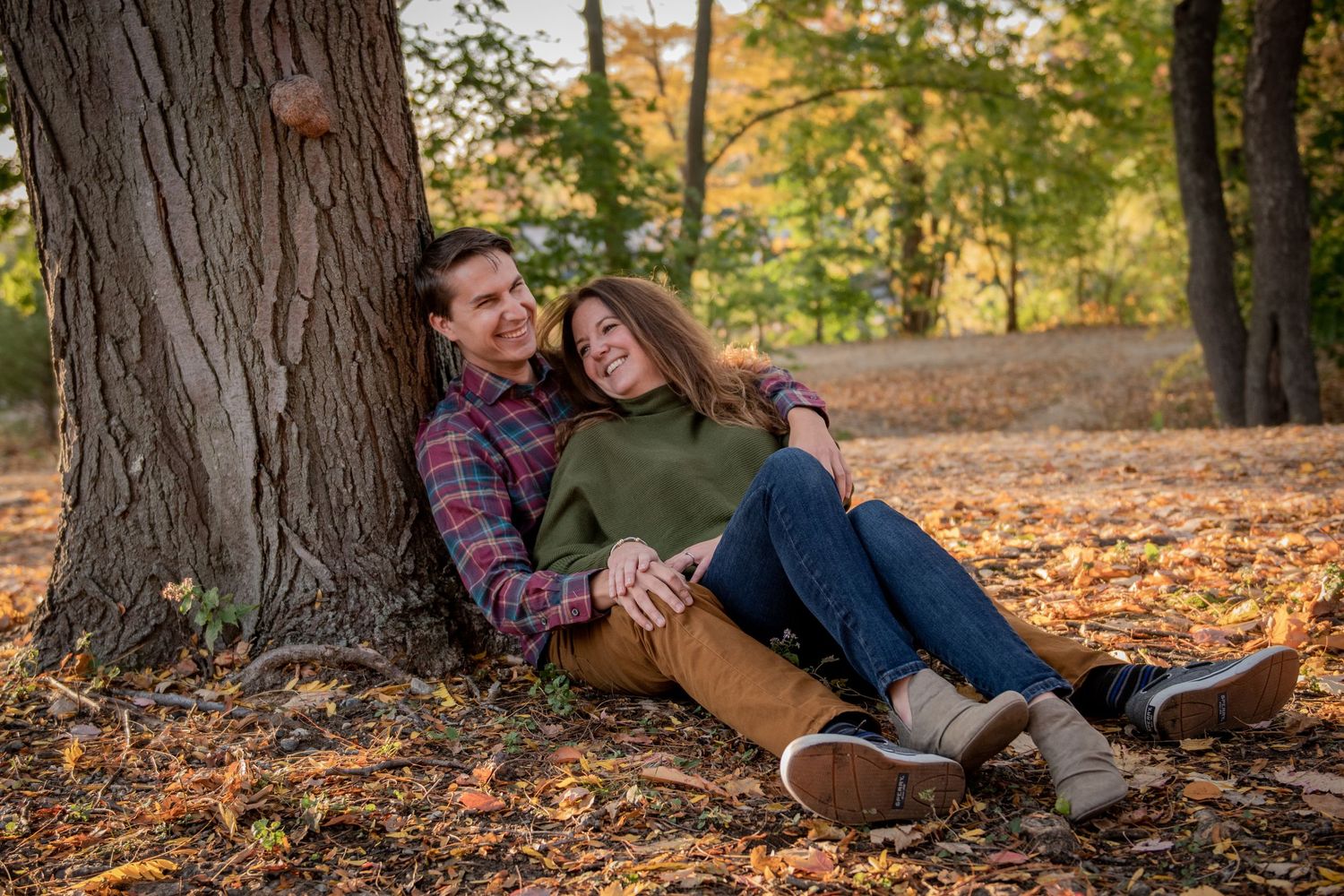 A couple relaxes against a large tree trunk surrounded by fallen autumn leaves. Massachusetts Engagement Photographer Serving New England