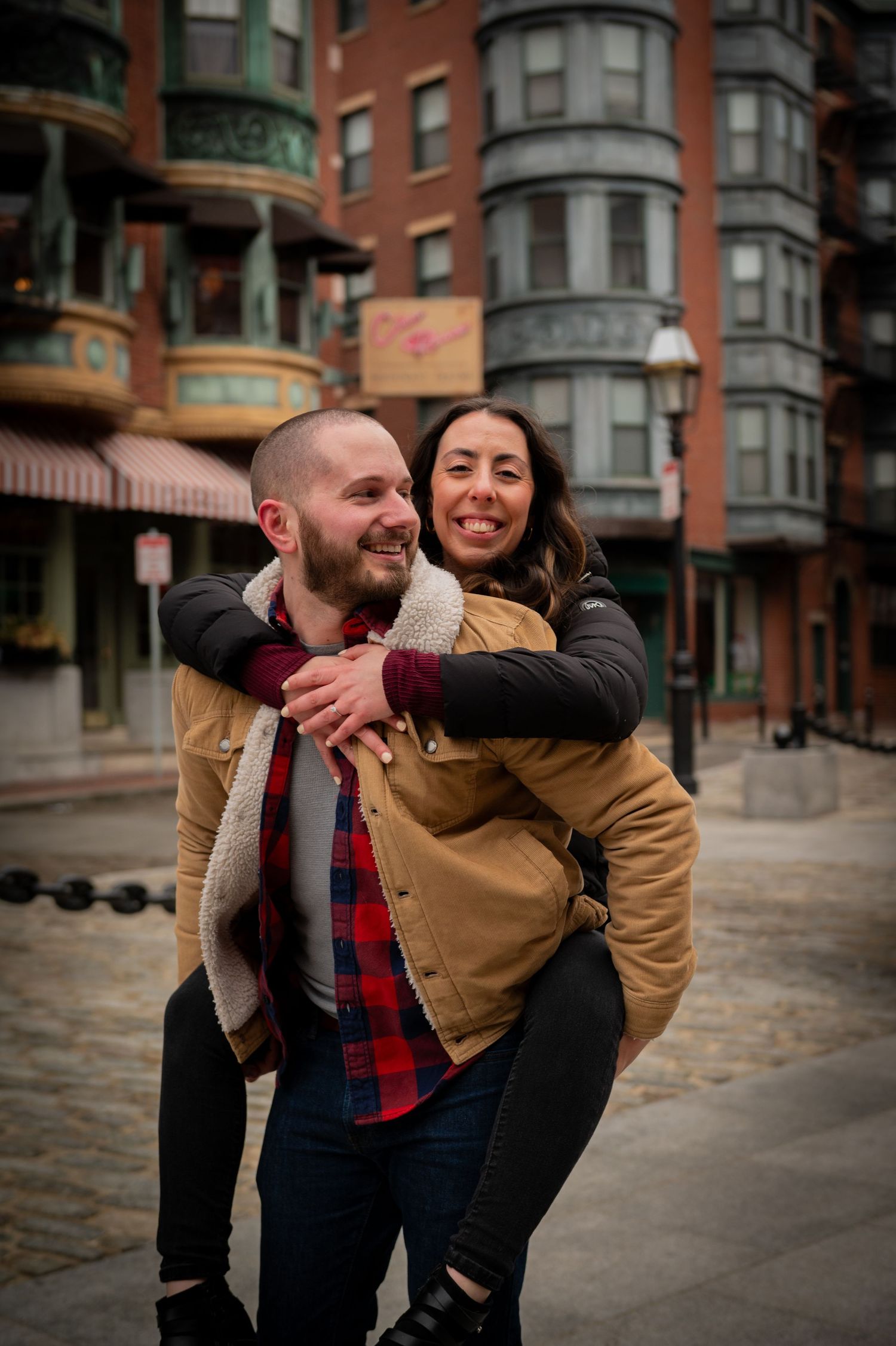 Playful piggyback ride moment against historic Boston brownstone buildings. Boston Engagement Photos- Affordable