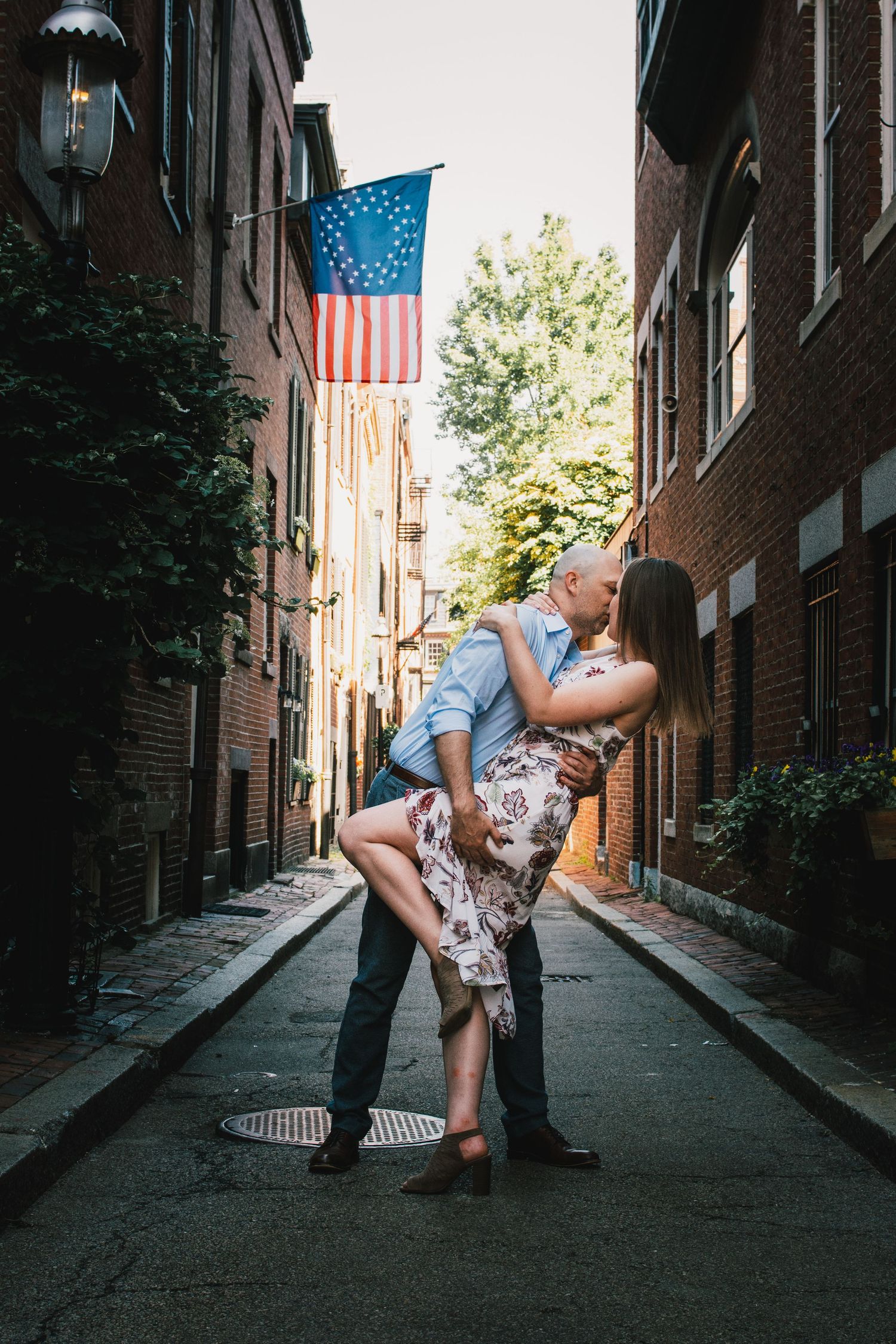 Couple sharing romantic moment in cobblestone alley with American flag overhead. Massachusetts Engagement Photographer Serving New England