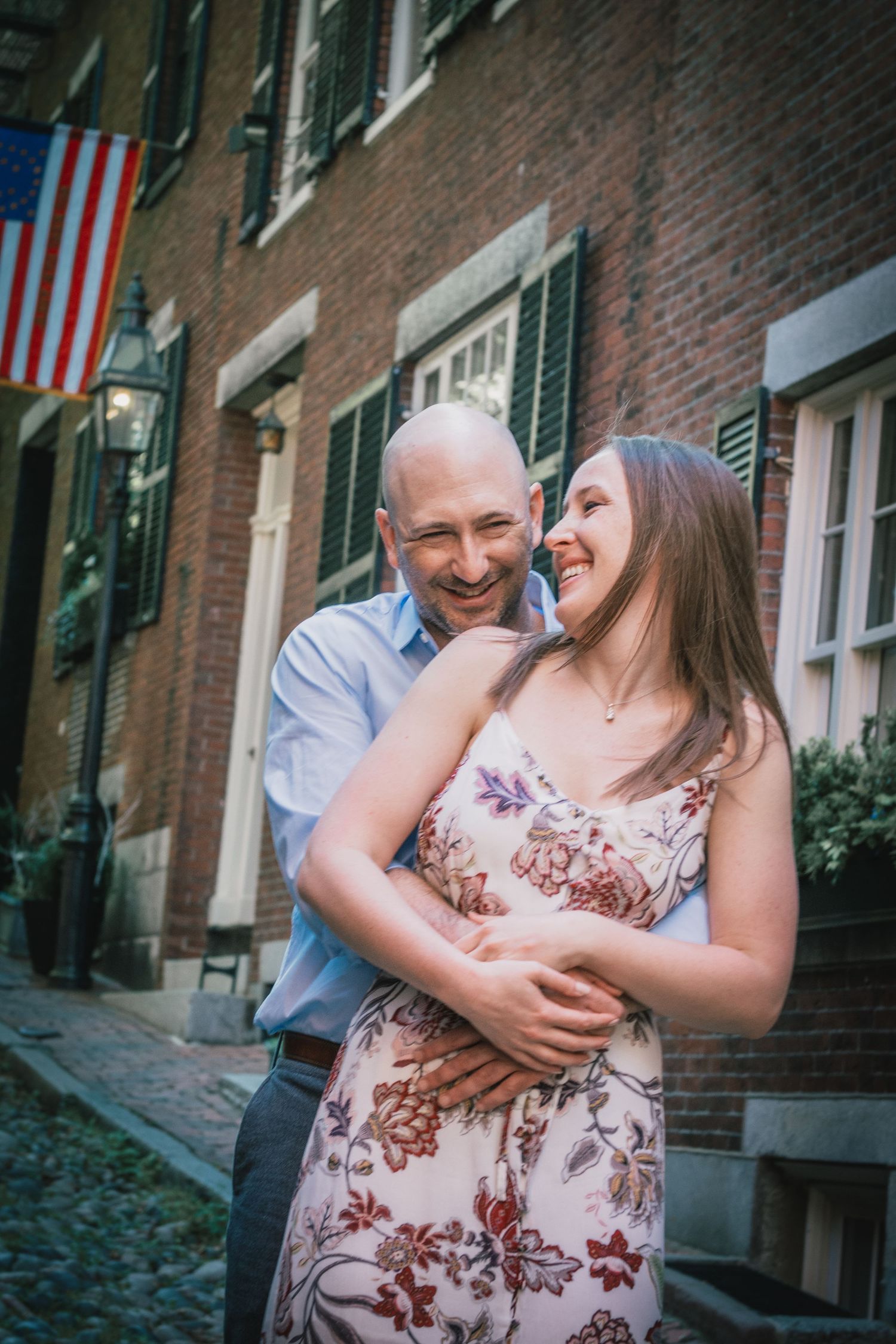 Happy couple embracing in front of historic brick building with American flag. Massachusetts Engagement Photographer Serving New England