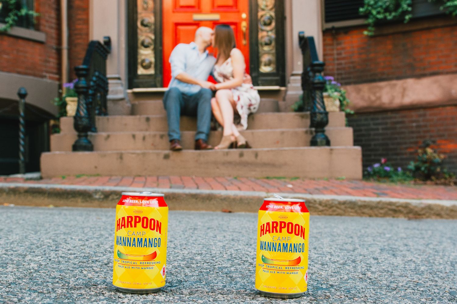 Two cans of Harpoon beer on street with couple sitting on steps in background. Massachusetts Engagement Photographer Serving New England