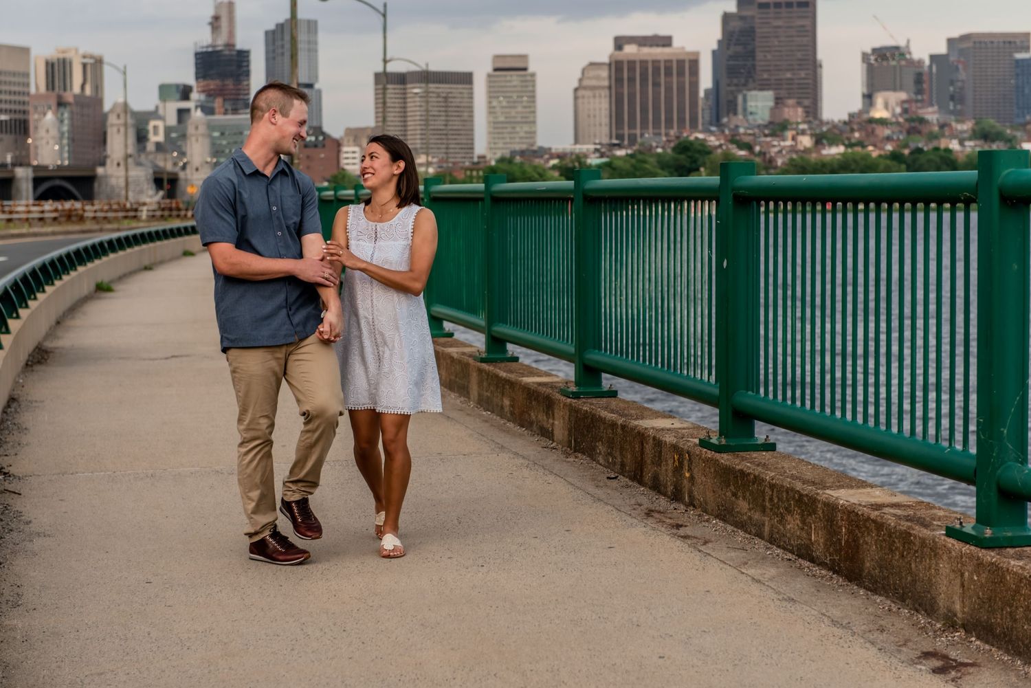 Couple walking along green bridge railing with city skyline in background. Massachusetts Engagement Photographer- Affordable