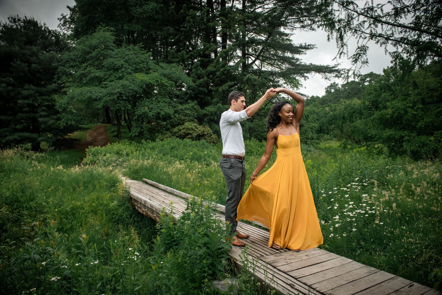 A couple dancing together on a wooden boardwalk surrounded by lush greenery. Massachusetts Engagement Photographer- Affordable