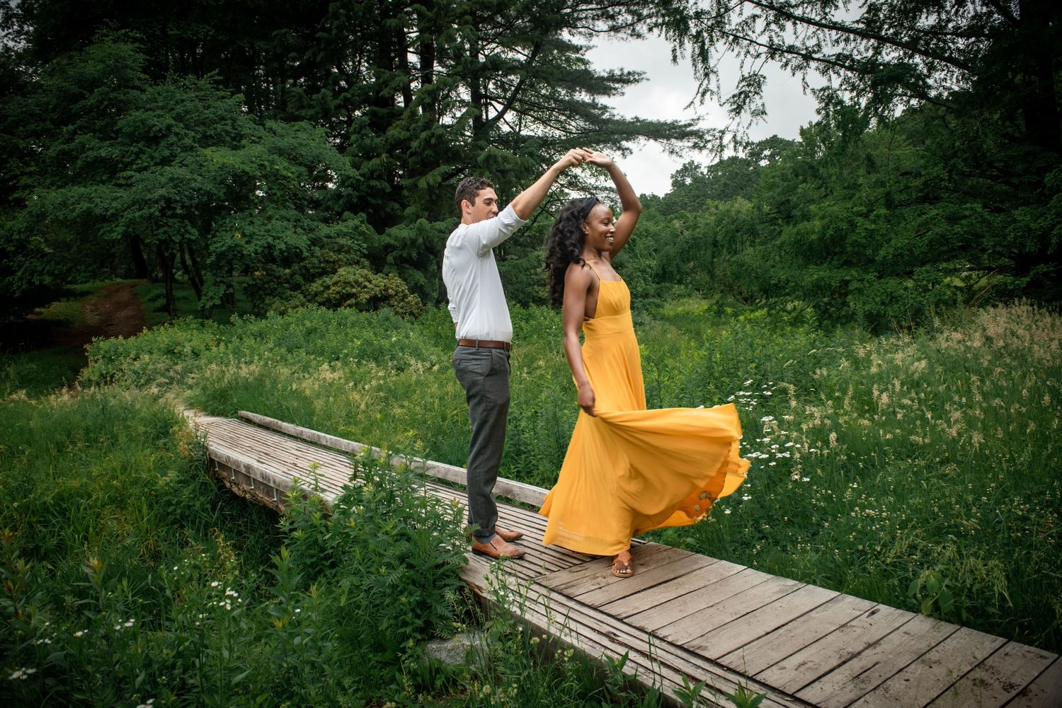 A couple twirling in a dance on a wooden walkway amidst wildflowers and trees. Massachusetts Engagement Photographer- Affordable