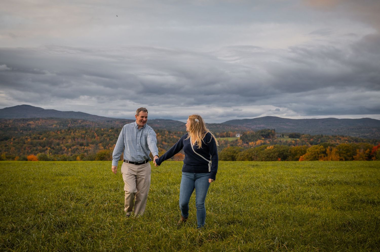 A couple walking hand in hand through an autumn field with mountains in the distance. New Hampshire Engagement Photos- Affordable
