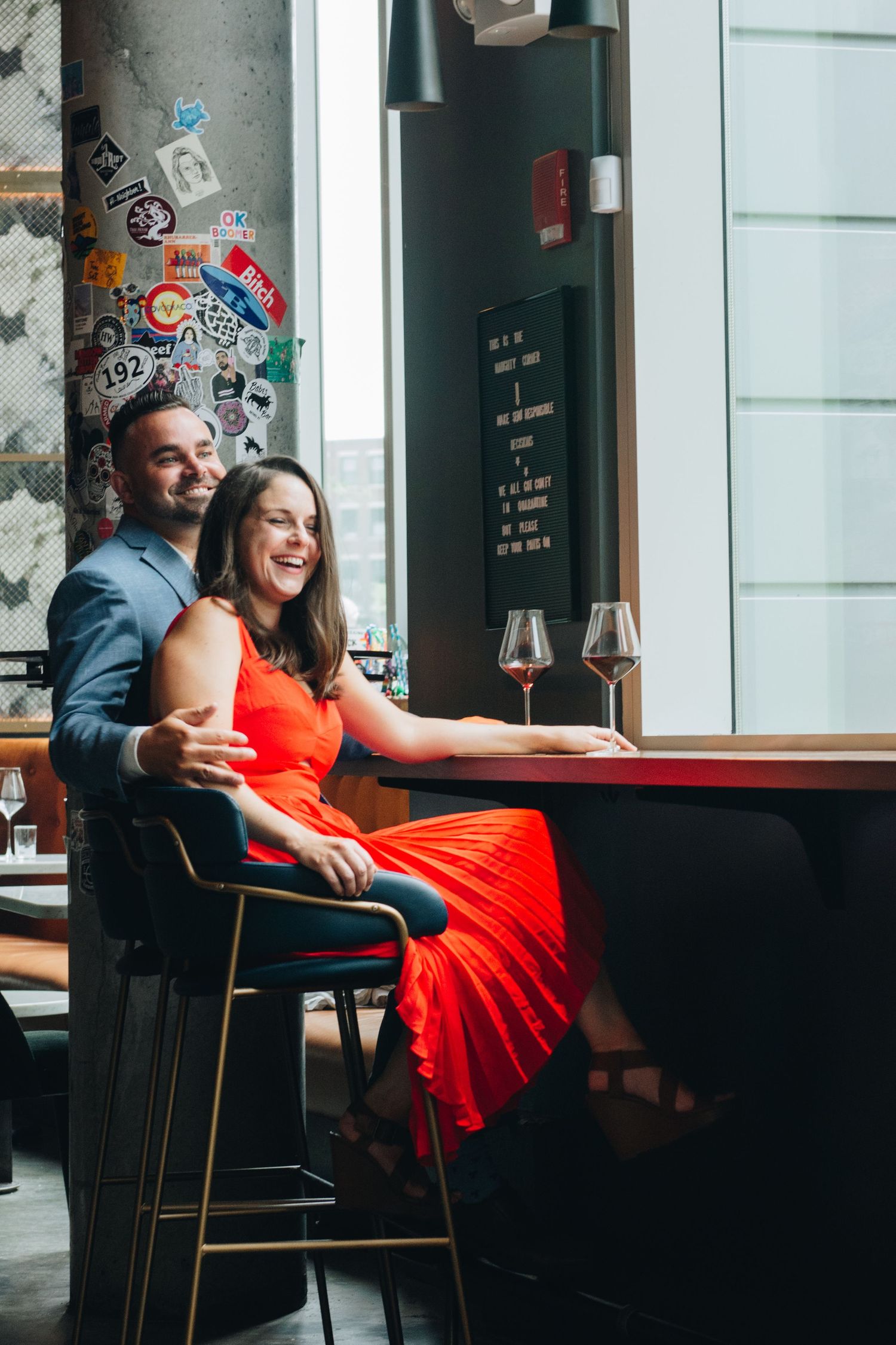 Couple enjoying wine at industrial-style bar with sticker-covered wall. Massachusetts Engagement Photographer Serving New England