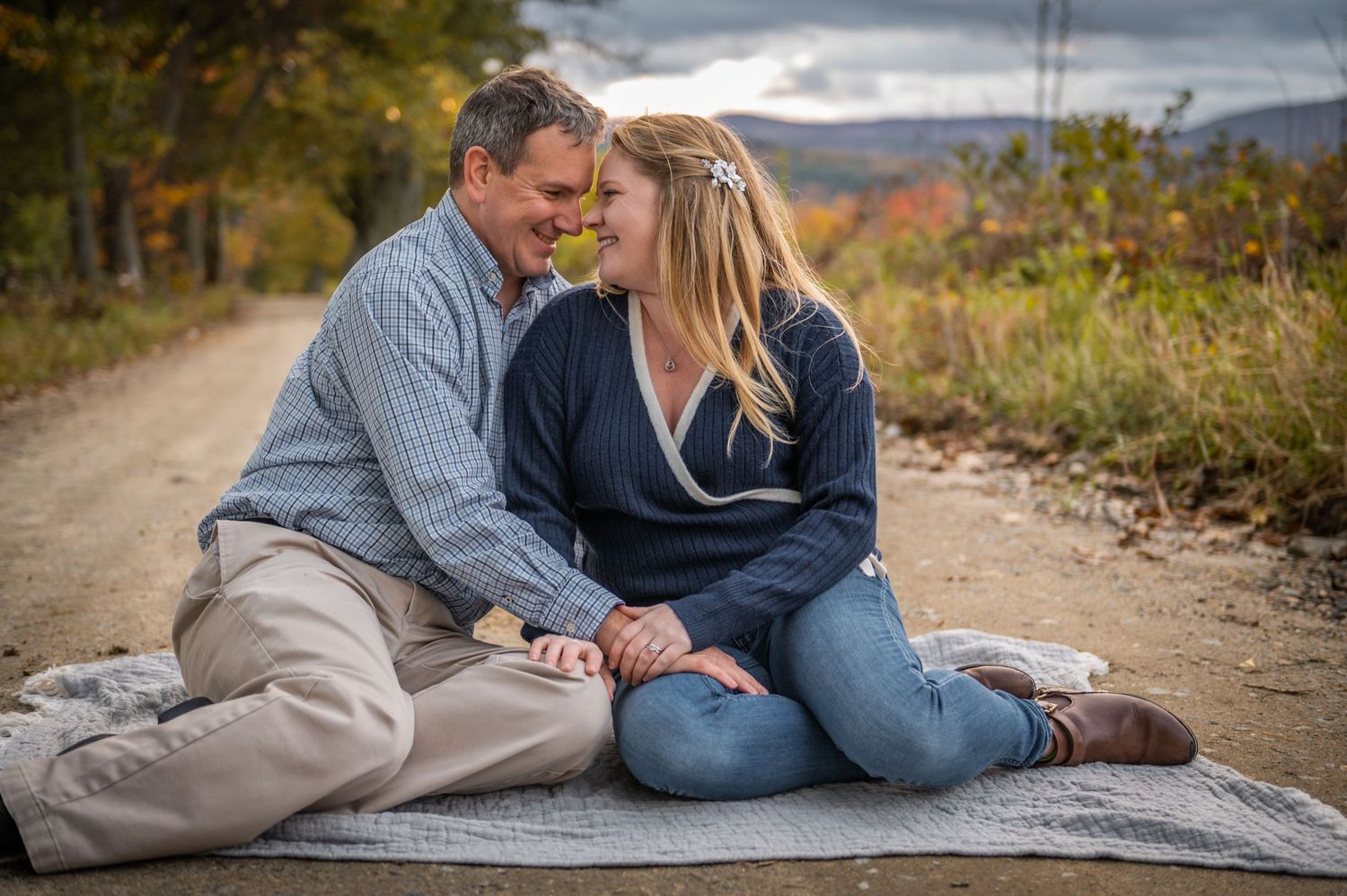 A couple sitting close together on a blanket on a dirt road surrounded by fall foliage. New Hampshire Engagement Photographer- Affordable