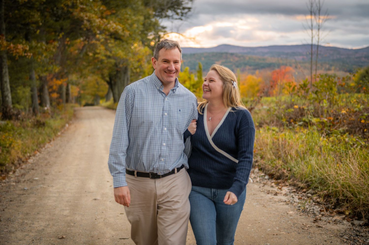 A couple walking along a scenic dirt road with fall foliage and mountains in the background. New Hampshire Engagement Photographer- Afforda