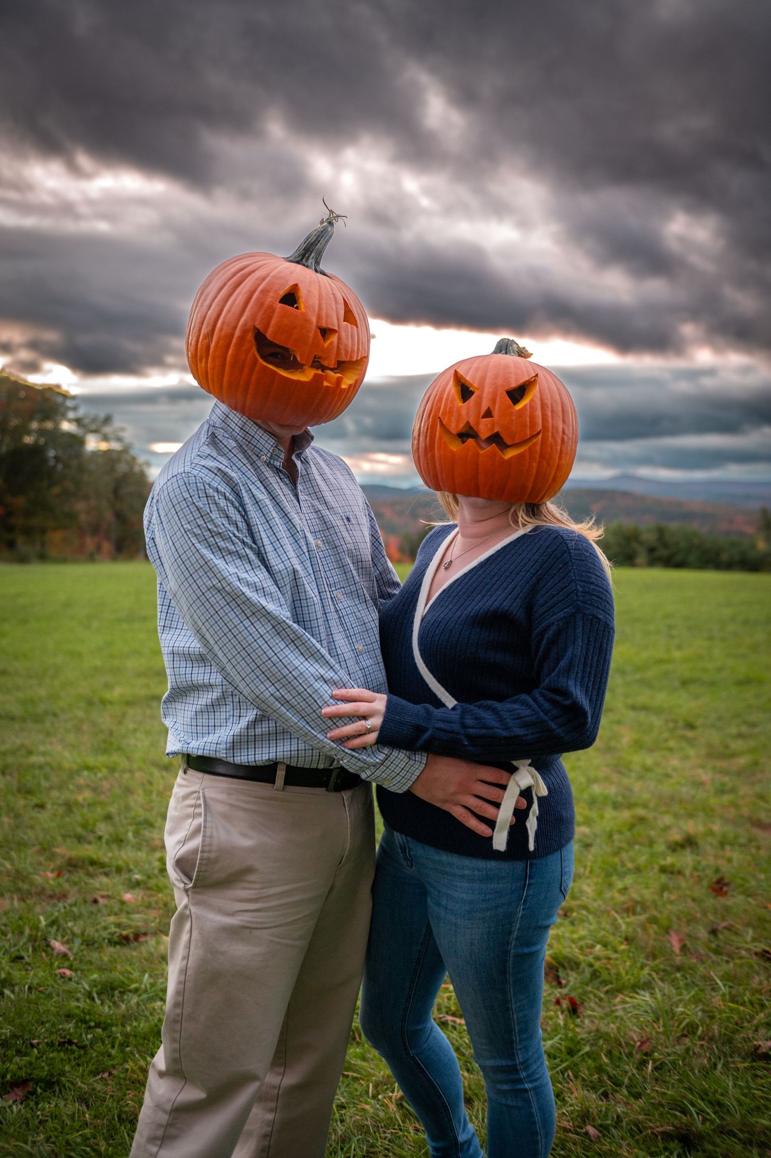 A couple wearing jack-o-lantern pumpkins on their heads pose in a field under dramatic clouds. New Hampshire Engagement Photographer
