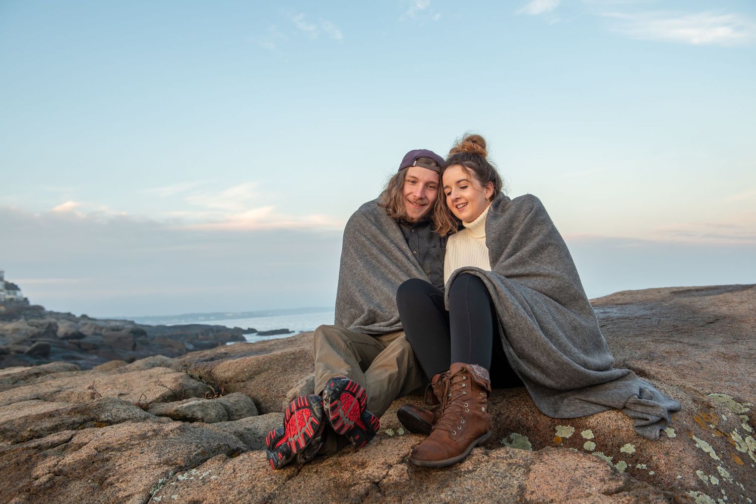 A couple wrapped in blankets sitting on coastal rocks at sunset.