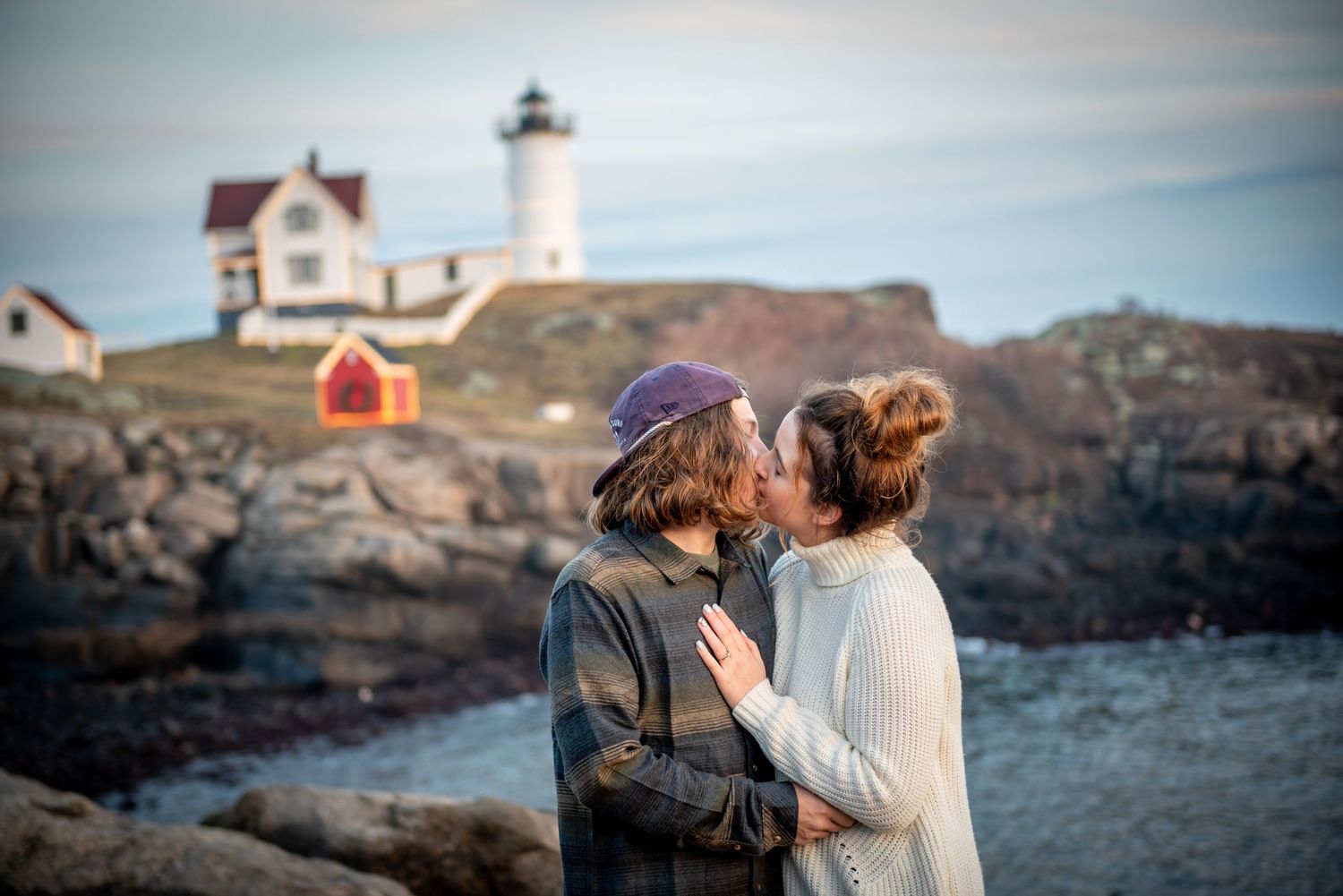 A couple shares a romantic moment in front of the iconic Nubble Lighthouse in Maine at sunset.