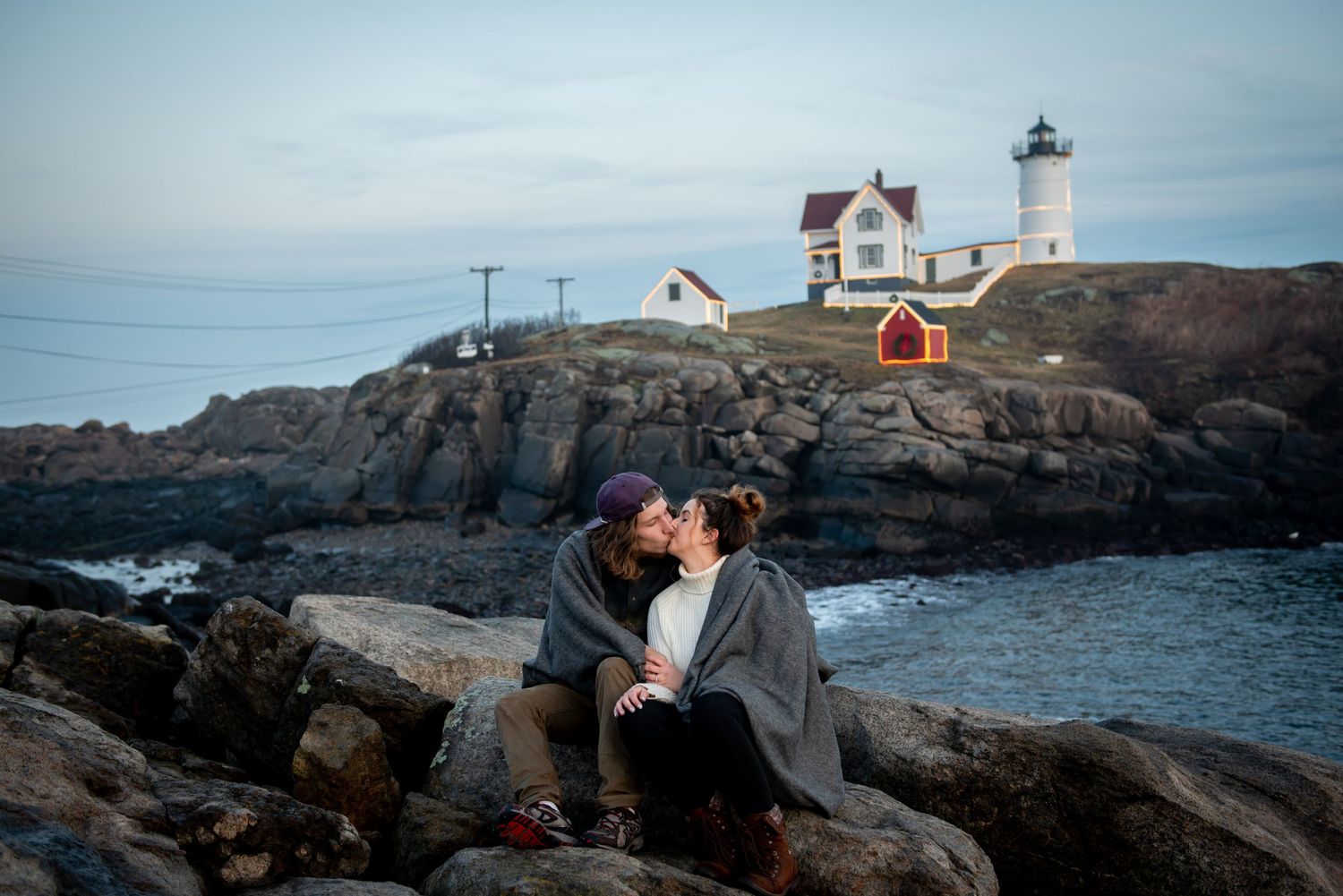 A romantic moment shared on rocky coastal cliffs with Nubble Lighthouse in the background. Maine Engagement Photographer Serving New England
