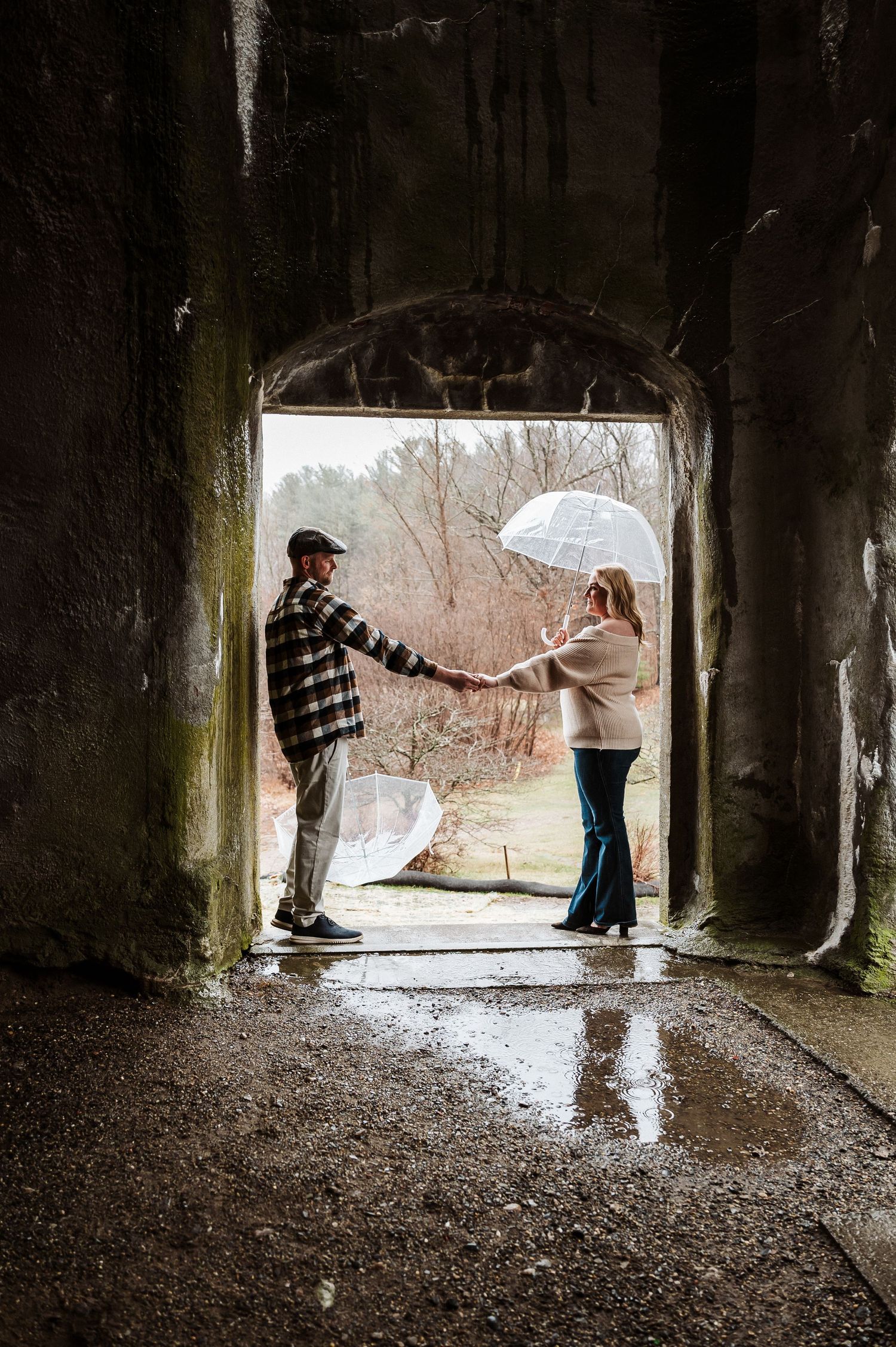 Two people stand in a stone archway during a rainy day, with one holding a white umbrella for shelter.