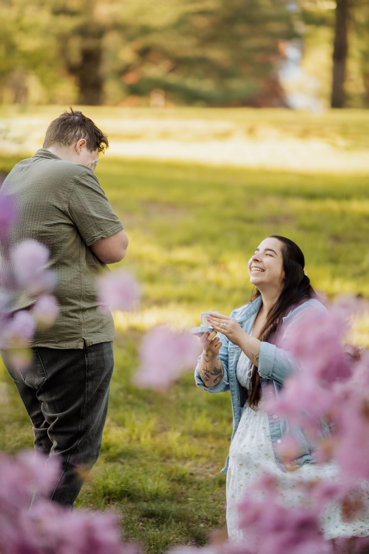 A marriage proposal takes place in a sun-dappled park with pink cherry blossoms framing the romantic moment.